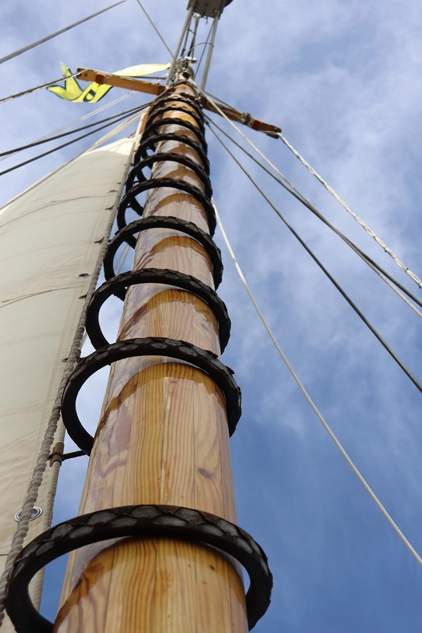 Looking up at the mast of a sailboat against a partly cloudy sky, showing the wooden mast, ropes, and rigging.