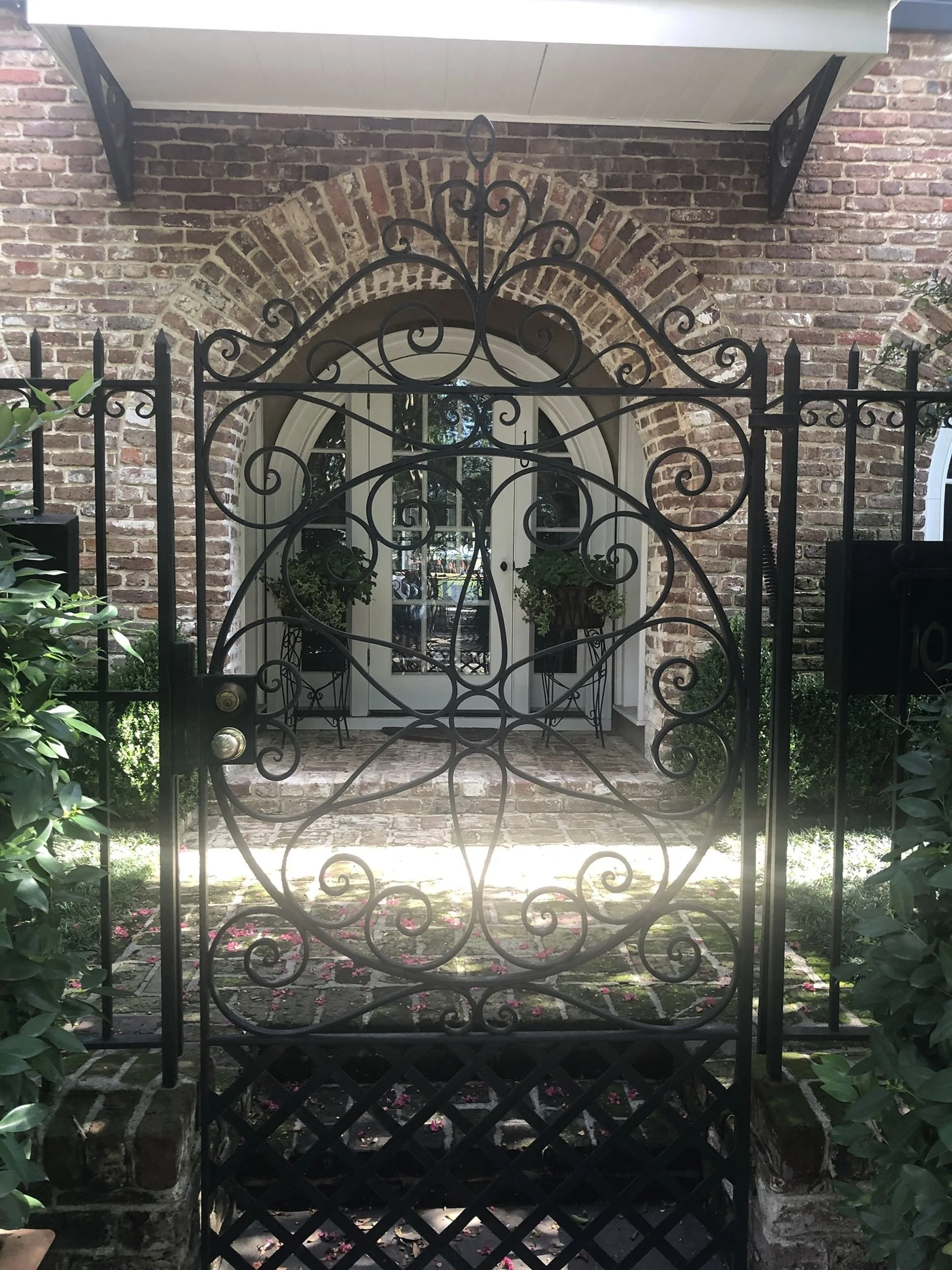 Through the Gate: Ornate wrought iron frames a private courtyard entrance, revealing layers of brick, greenery, and classic Charleston craftsmanship beyond. Rich in detail and visual intrigue, this image evokes exclusivity, elegance, and discovery—we