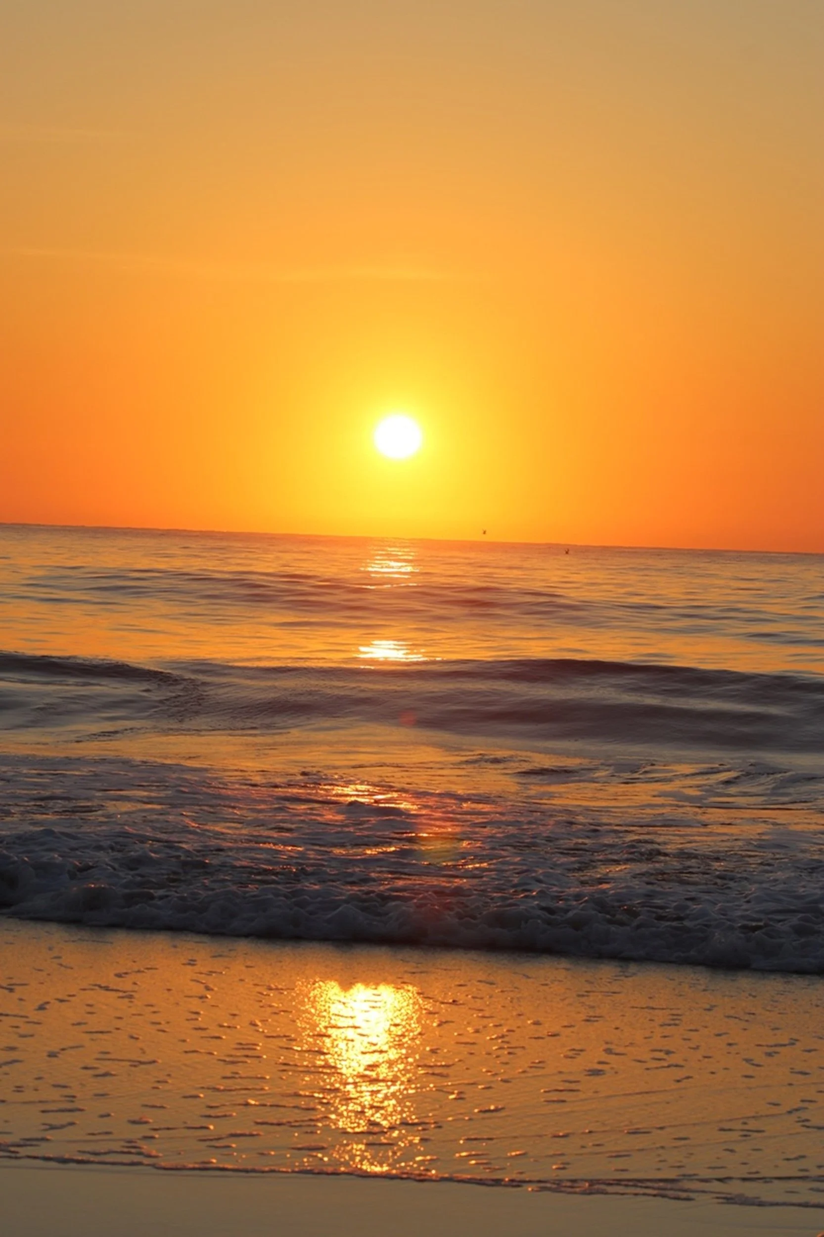 Sunset over the ocean with the sun reflecting on the water, gentle waves, and orange sky.