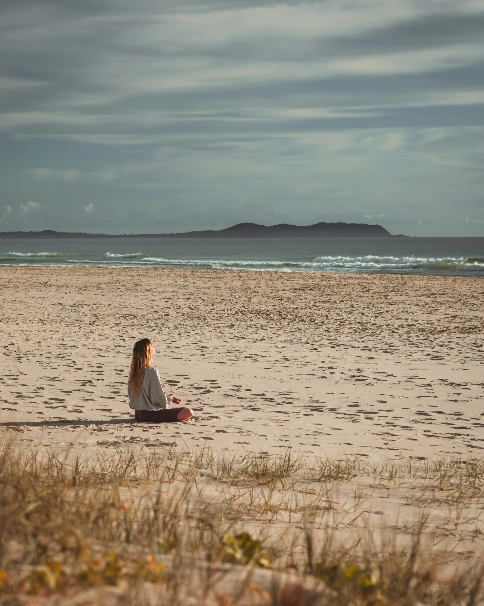 A woman with long hair sits cross-legged on the sandy beach facing the ocean. She is dressed in a beige sweatshirt and dark pants, meditating or relaxing. The beach has some sparse grass and plants in the foreground. The ocean waves gently hit the shore, and there's a distant landmass or island with hills in the background. The sky is cloudy with streaks of clouds.