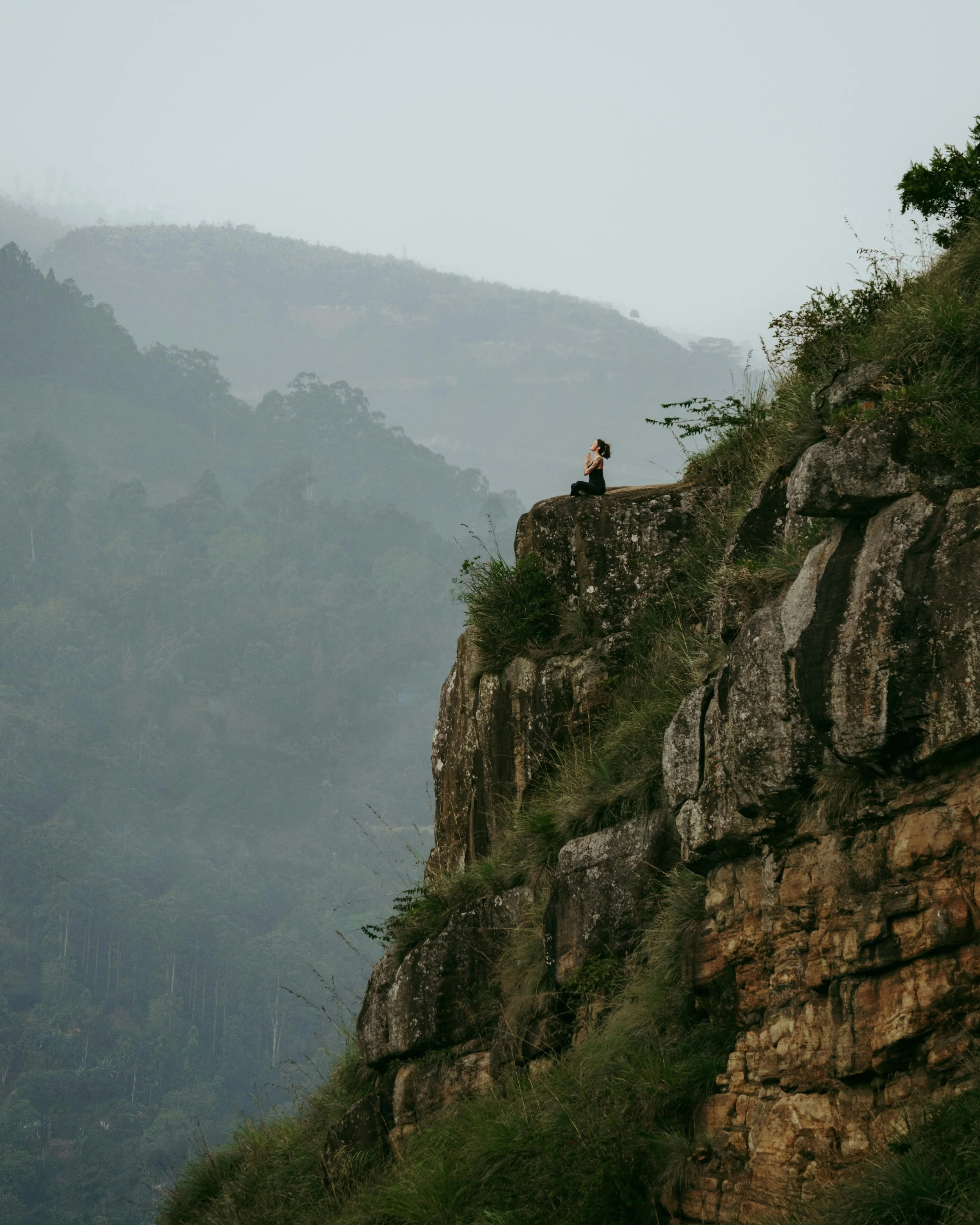 Person sitting on a large rock ledge on a hillside, meditating with hands in prayer position, overlooking a misty, green mountain landscape.