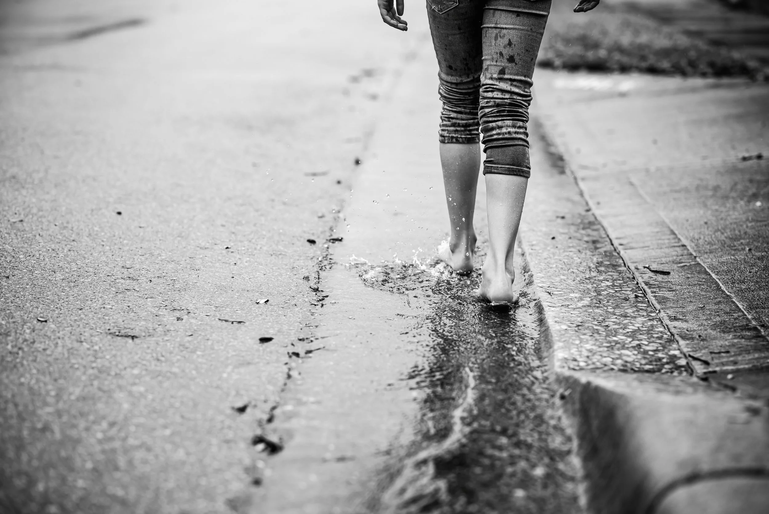 A person walking barefoot on a wet sidewalk, with water splashing around their feet, wearing rolled-up jeans.