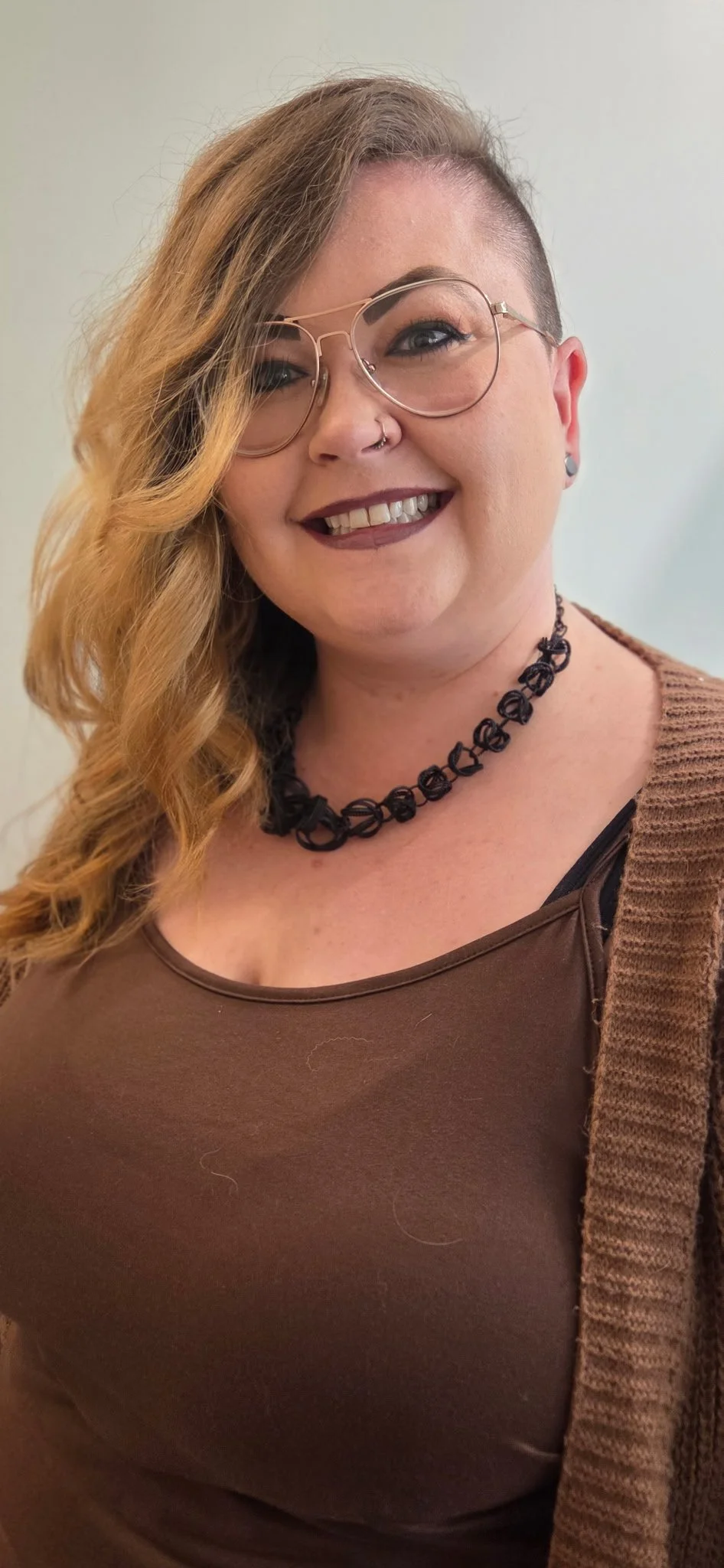 A woman with dyed blonde and brown hair, wearing glasses, a black choker, a brown top, and a brown cardigan, smiling at the camera.