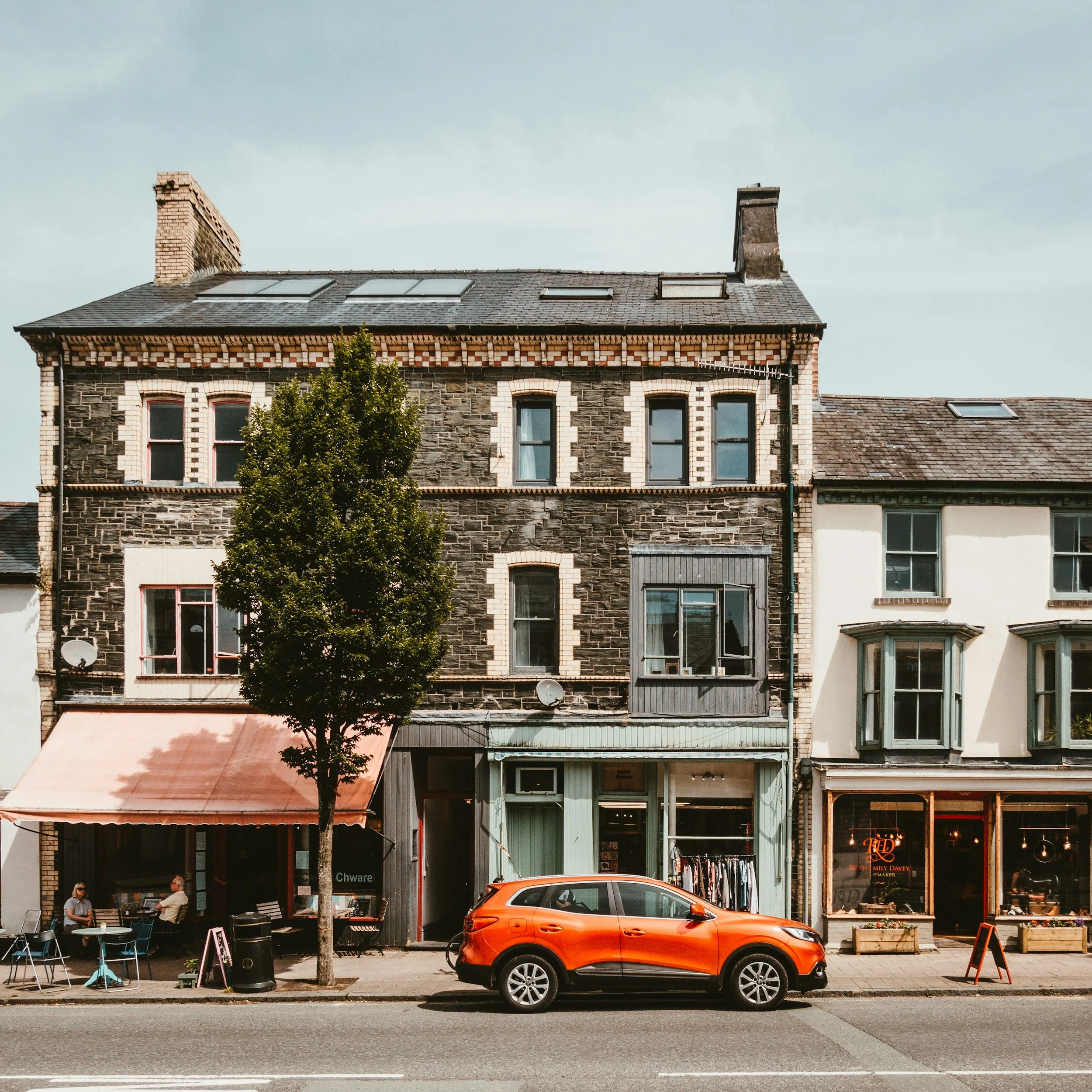 A stree view featuring a mixed-use building with retail shops on the ground floor, a brown brick facade on the second floor, and various windows. In front, a bright orange SUV is parked on the street, with pedestrians sitting outside at a cafe under a pink awning, a tree, and a sidewalk.