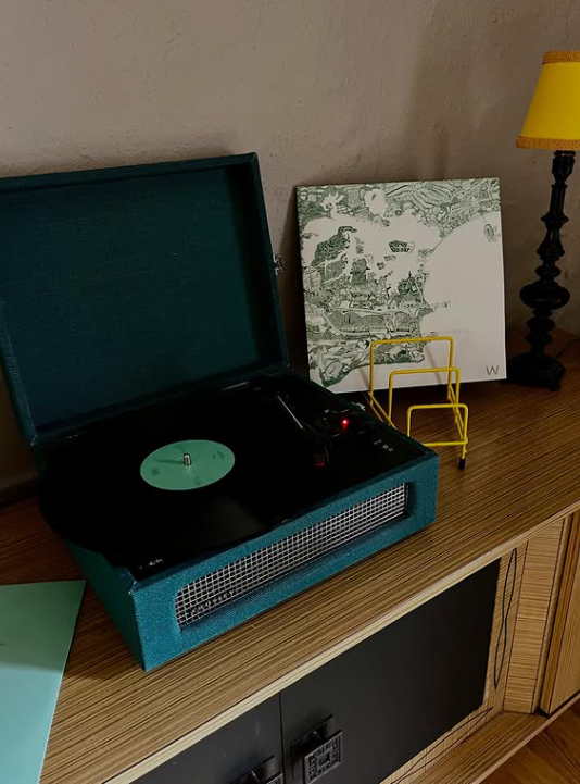 A vintage teal-colored portable record player with a black turntable, open lid, and spinning record on a wooden sideboard. Behind it, a black and white map art print is leaning against a wall, and to the right, a yellow and black table lamp is turned
