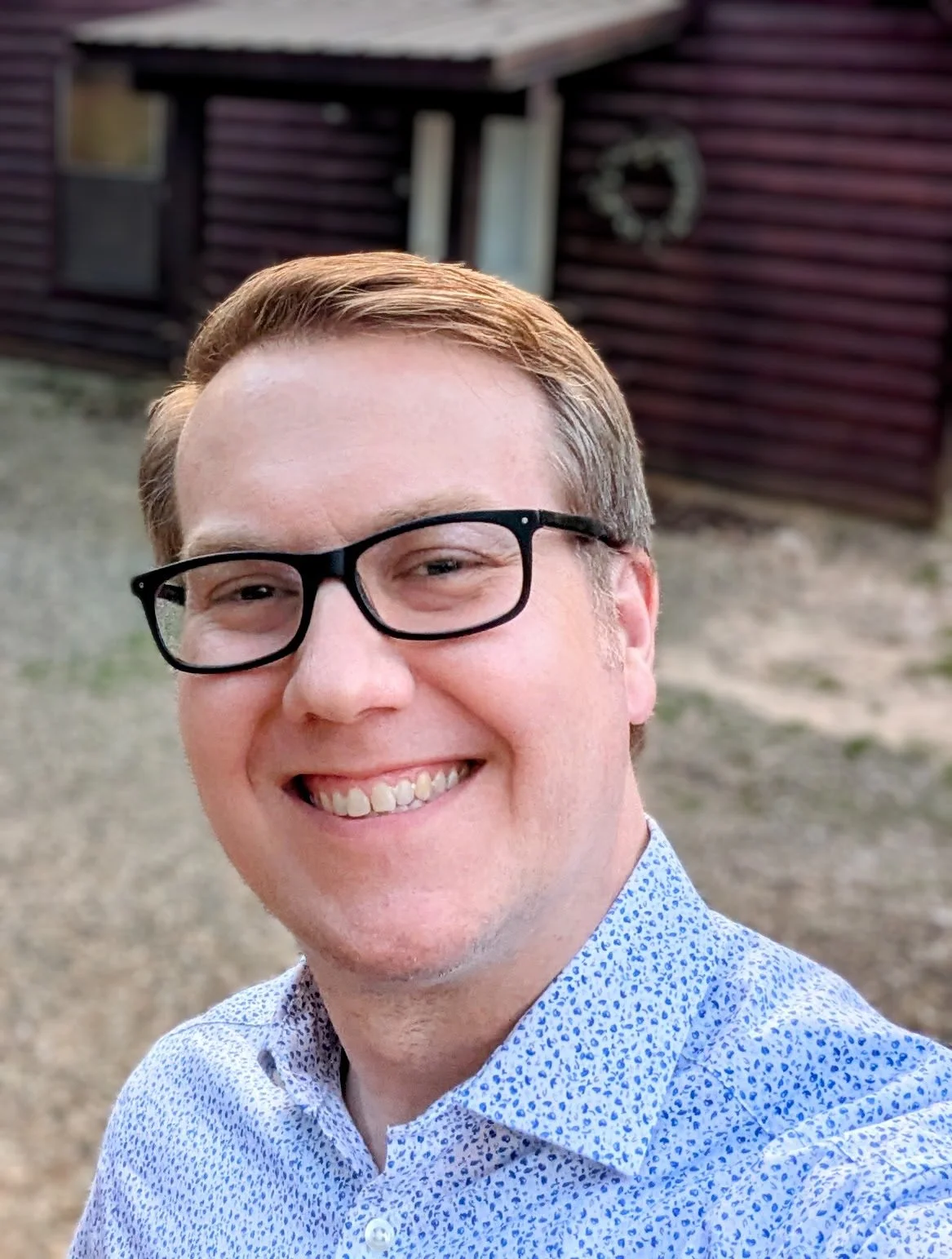 A smiling man with glasses and light brown hair taking a selfie outdoors in front of a dark wooden building.