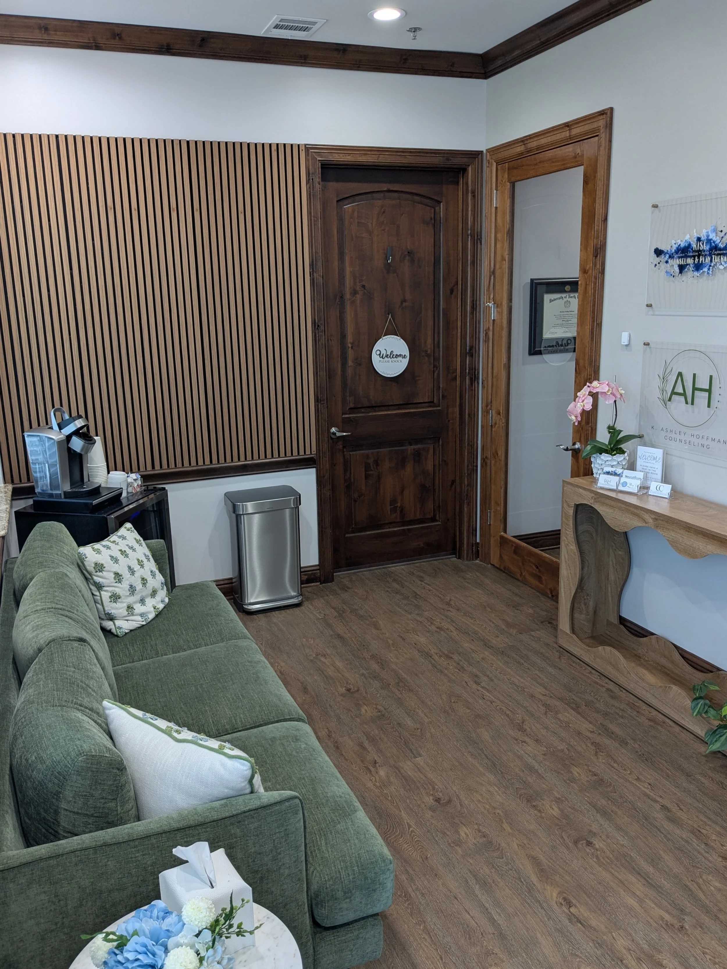 Interior of an office or waiting room with a green sofa, a small side table with flowers, a wood-paneled wall, a wooden door with a welcome sign, and a glass door with framed documents and decorations.