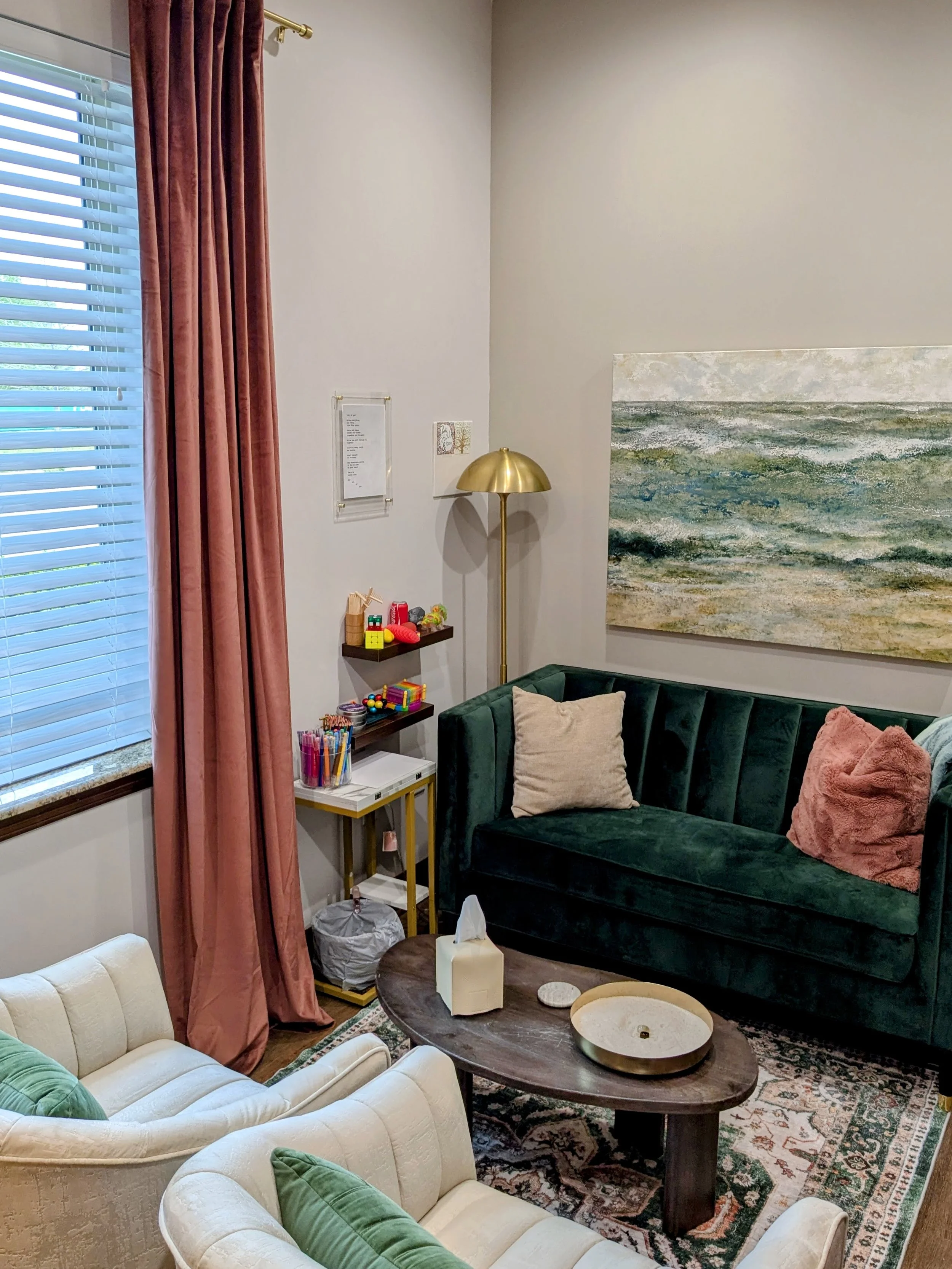 Living room with a green velvet sofa, white armchairs, and a wooden coffee table; window with pink curtains; abstract seascape painting on wall; gold floor lamp and small shelf with toys and art supplies.