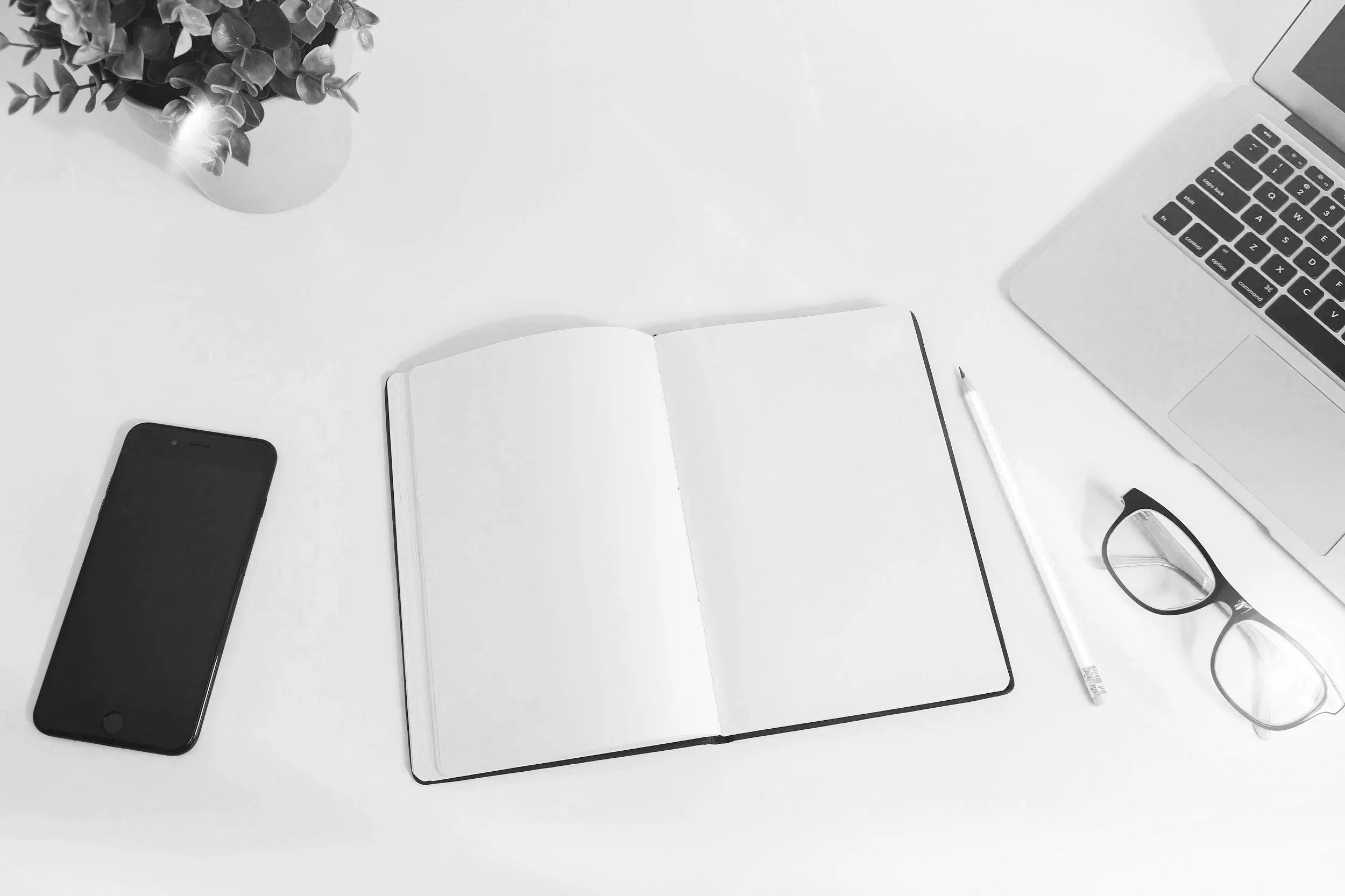 Mesa de trabajo con un cuaderno abierto, una tableta, un teléfono móvil, un bolígrafo, unas gafas y una laptop, con una planta en la esquina superior izquierda.