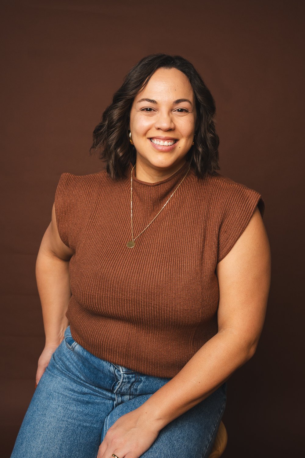 A woman with shoulder-length dark curly hair, smiling, wearing a rust-colored sleeveless top and blue jeans, sitting against a brown background.