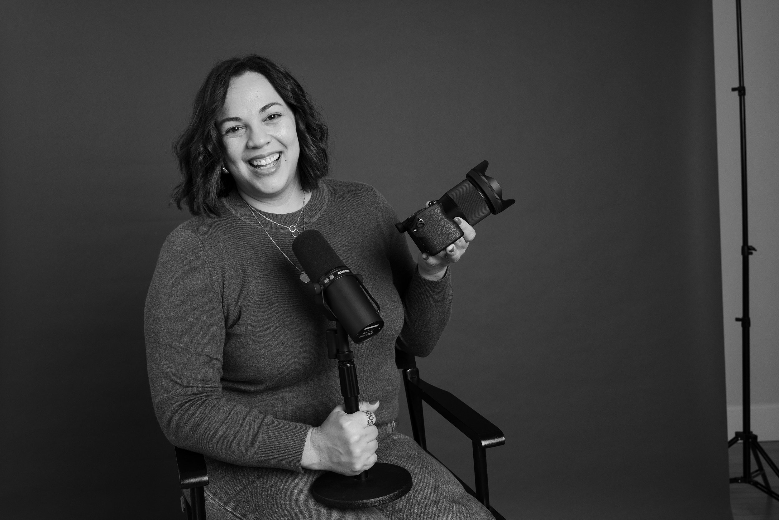 Mujer con cabello rizado y sonrisa, sosteniendo cámara y micrófono en un estudio con fondo oscuro.