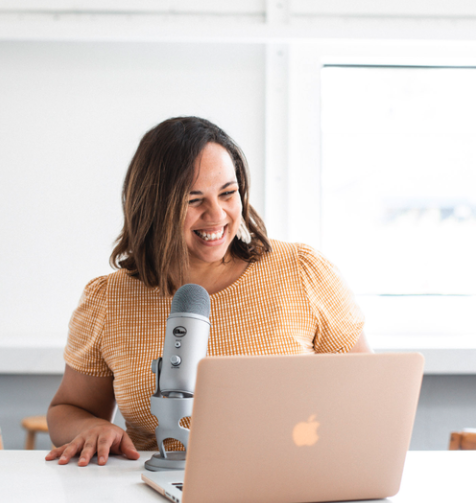 A woman with shoulder-length brown hair sitting at a desk, smiling at her laptop with a microphone nearby, in a bright room with white walls and a window in the background.