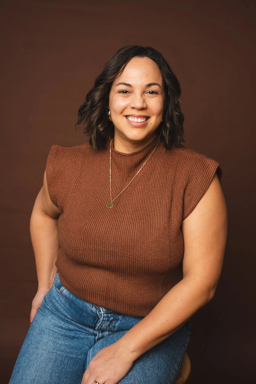 Retrato de una mujer sonriendo, con cabello rizado y oscuro, lleva una camiseta marrón y jeans azules, con un fondo marrón oscuro.