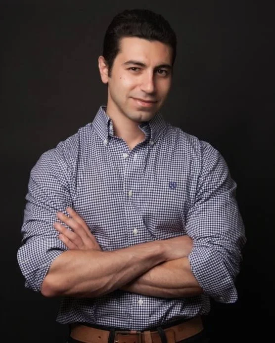 A young man with dark hair, wearing a checkered button-up shirt, standing with arms crossed against a dark background, smiling slightly.