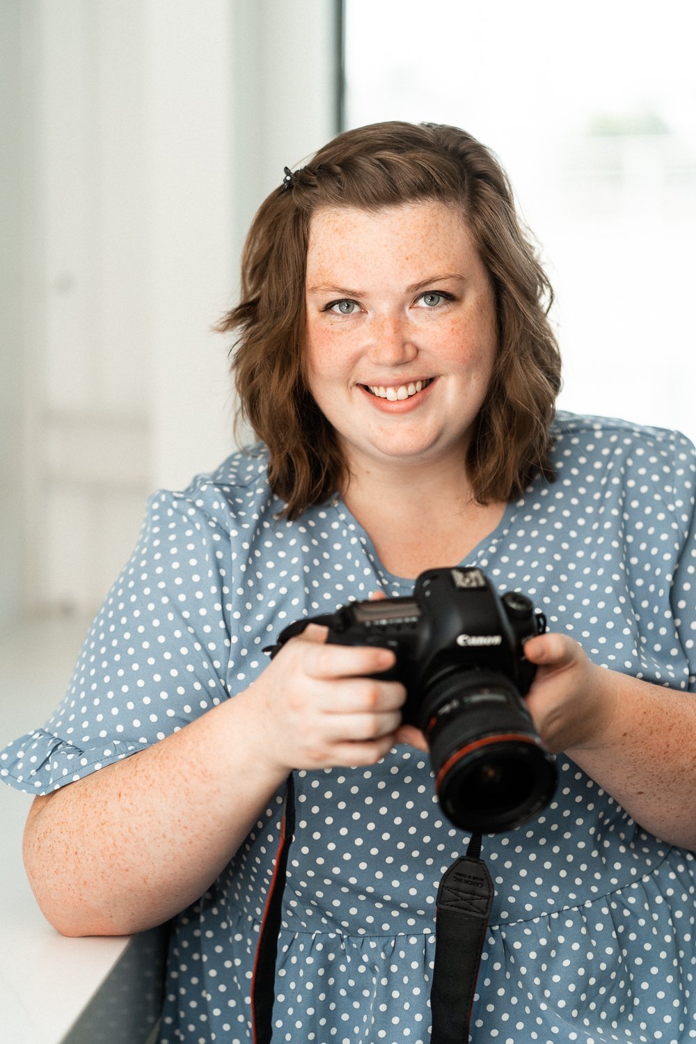 A smiling woman with wavy brown hair holding a Canon camera.