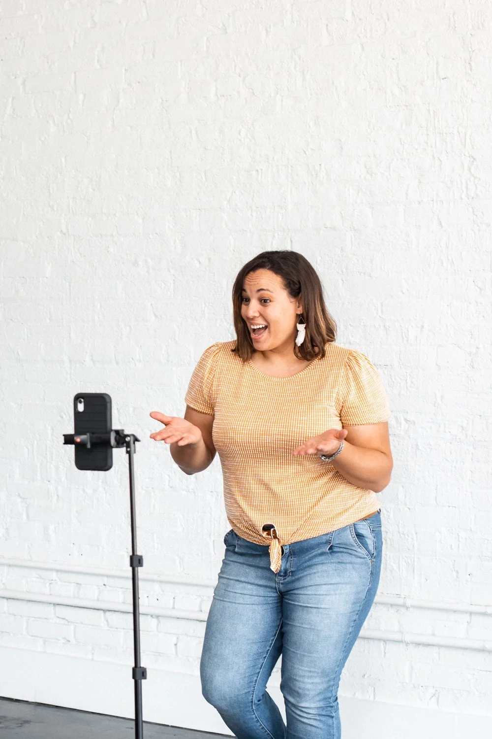 A woman standing in front of a white brick wall, looking excited and gesturing towards a smartphone on a tripod, as if recording a video.