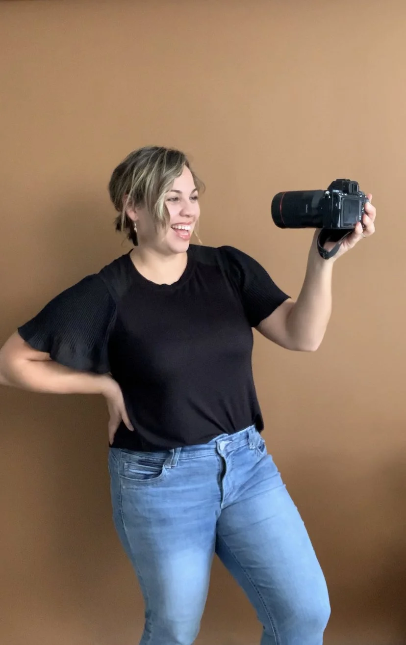 A woman smiling and holding a camera in front of a plain brown wall.