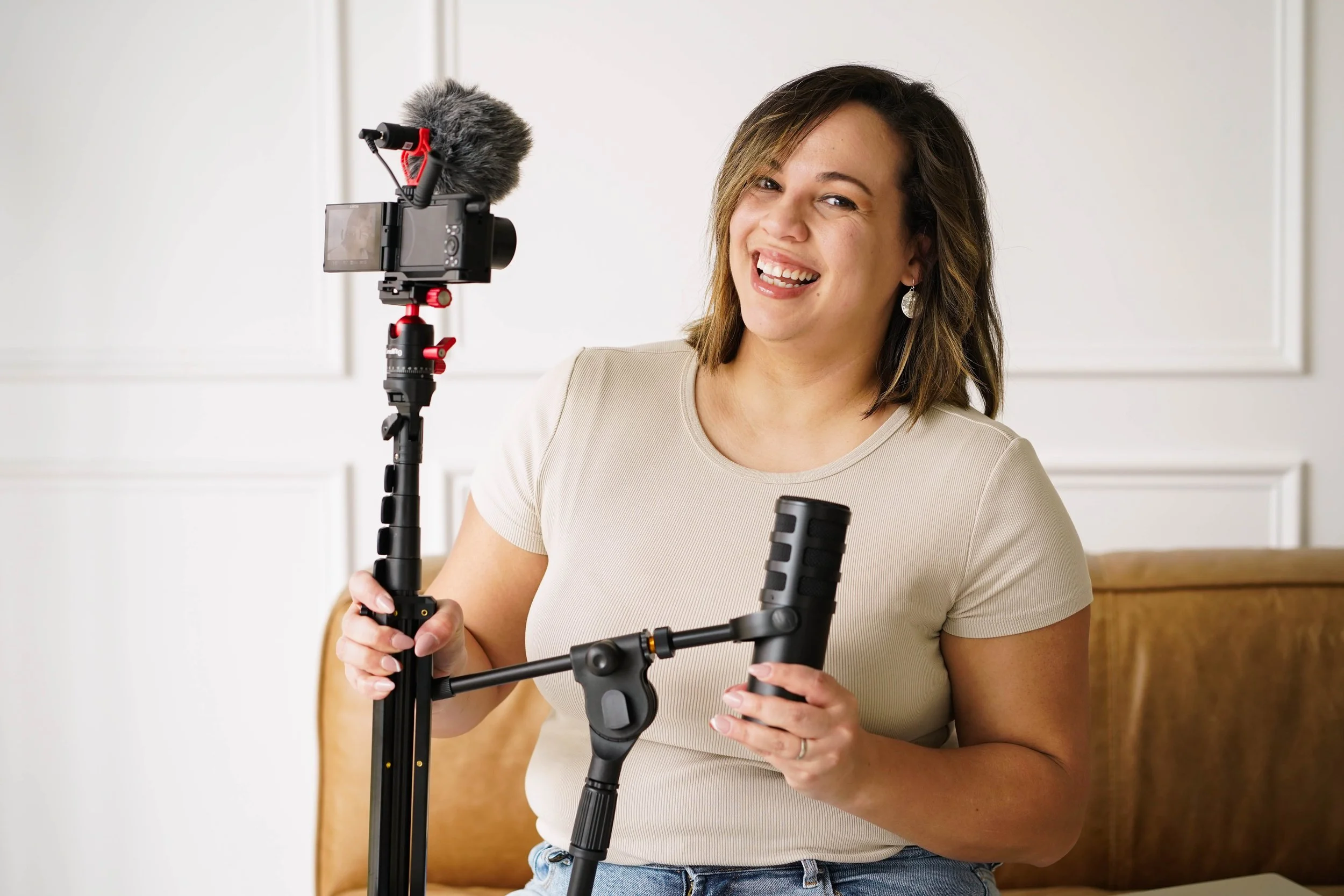 Woman holding a camera stabilizer with a mounted microphone and a camera, smiling in a casual indoor setting.