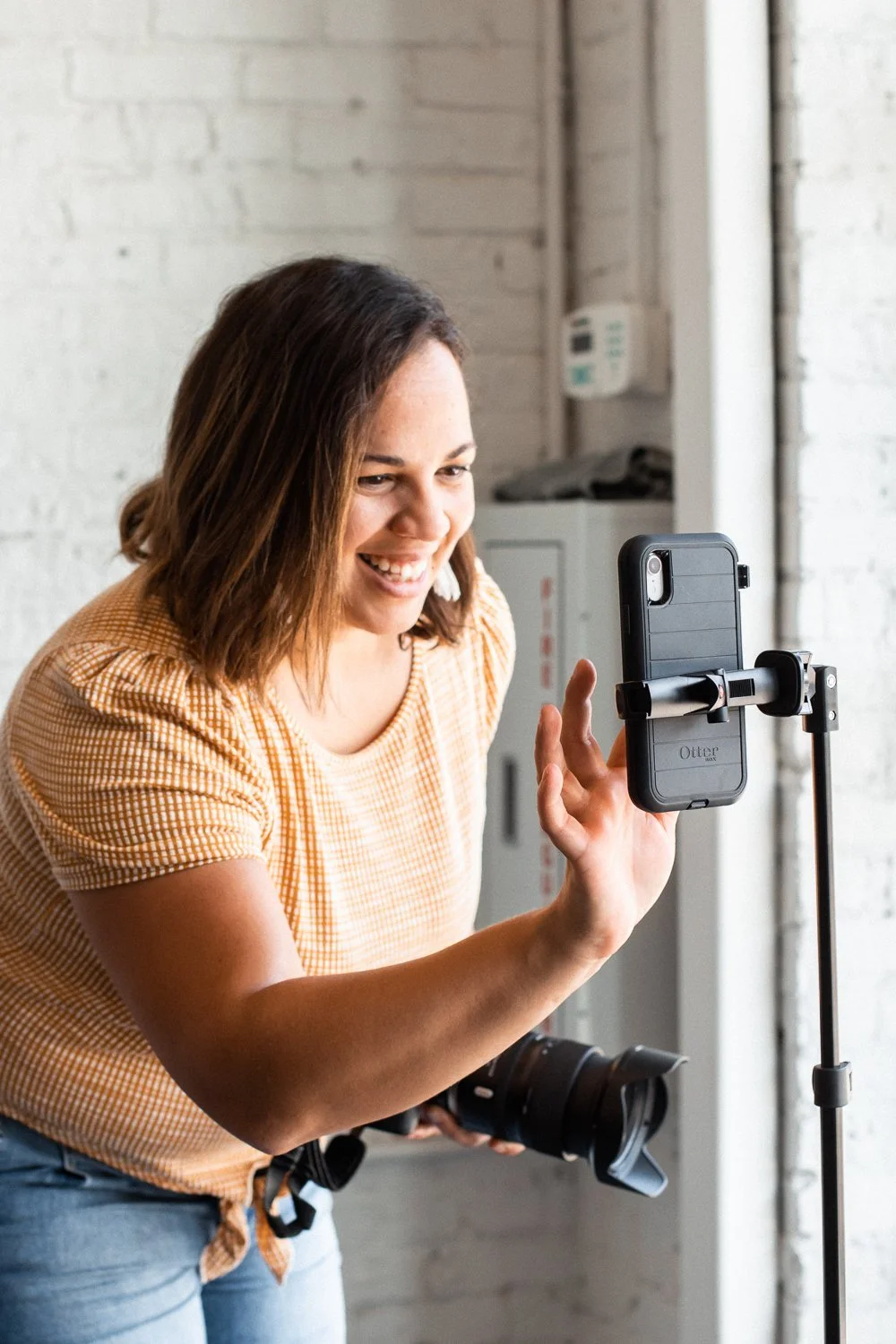 Una mujer sostiene un teléfono en un soporte, sonriendo y tomándose una selfie en un entorno interior con paredes de ladrillo blancas y equipo de oficina visible.