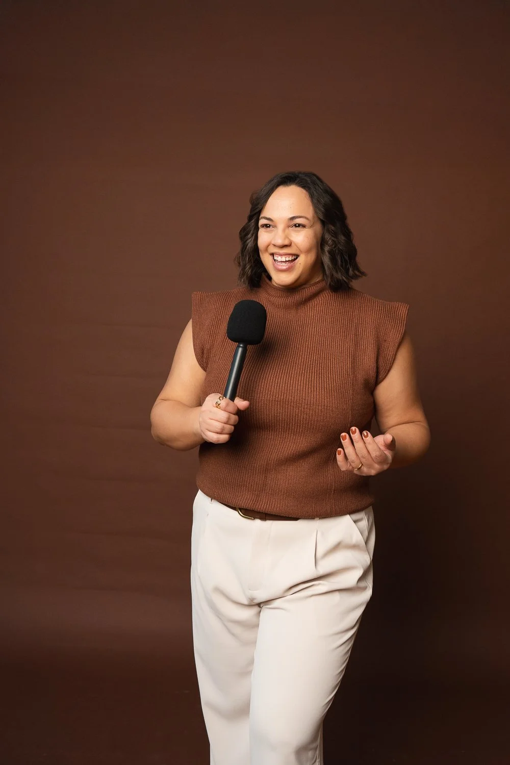 Mujer sonriente sosteniendo un micrófono en un fondo marrón.