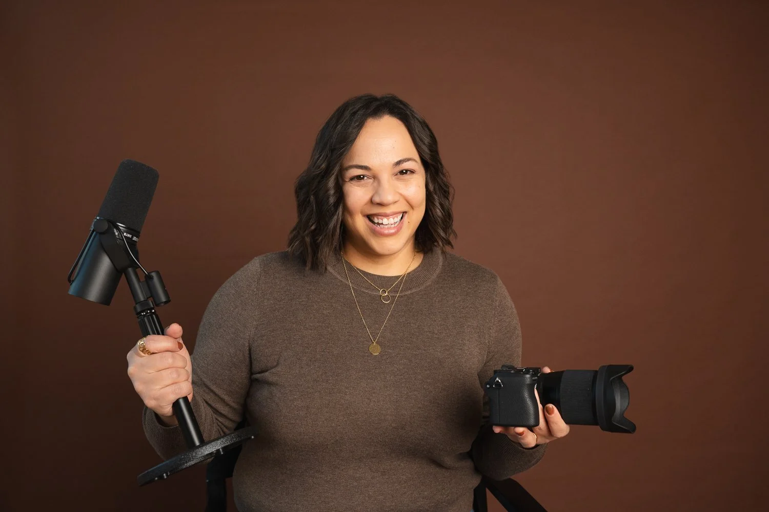Mujer sonriente sosteniendo una cámara y un micrófono en un fondo marrón.