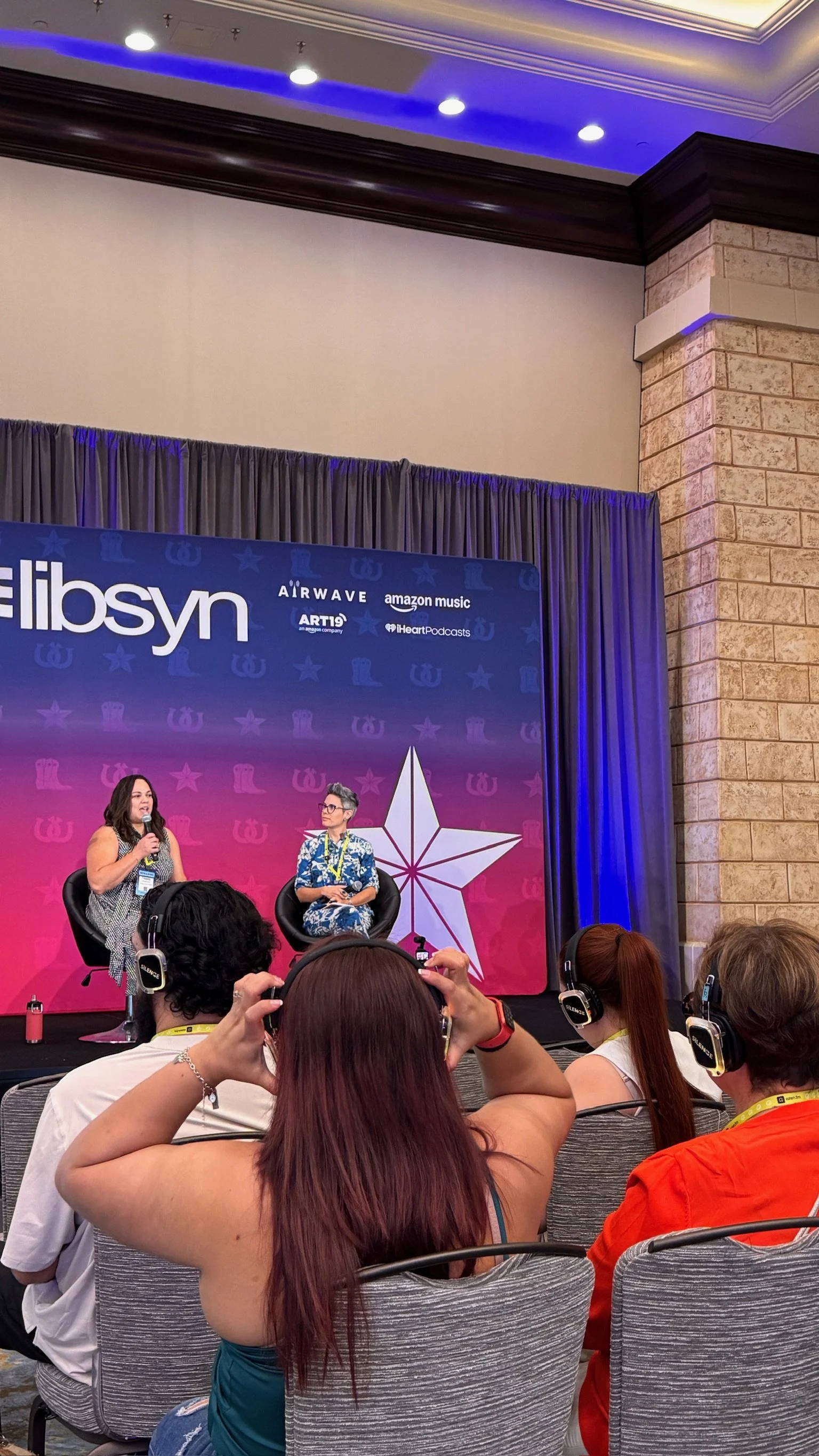 Evento en escenario con fondo azul y rosa, con logo de Allyson y patrocinadores como Amazon Music, Airwave, ART19 y iHeart Podcasts. Dos mujeres en la entrevista, una con micrófono, y audiencia con audífonos.