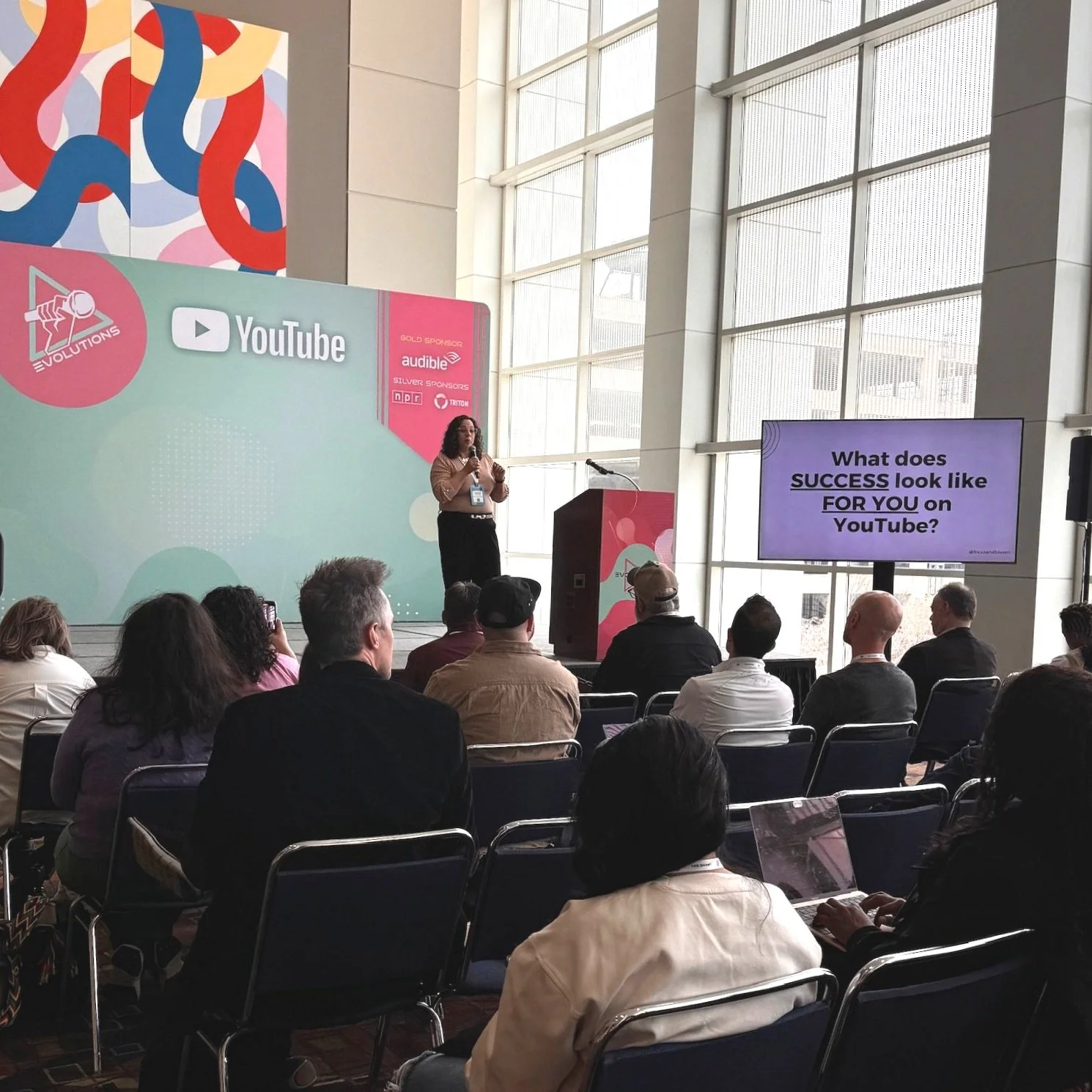 Una mujer presenta en un escenario con un fondo que tiene el logotipo de YouTube y patrocinadores. La audiencia observa y algunos toman notas o usan sus computadoras. En una pantalla se lee la pregunta: ¿Qué aspecto tiene el éxito para ti en YouTube?