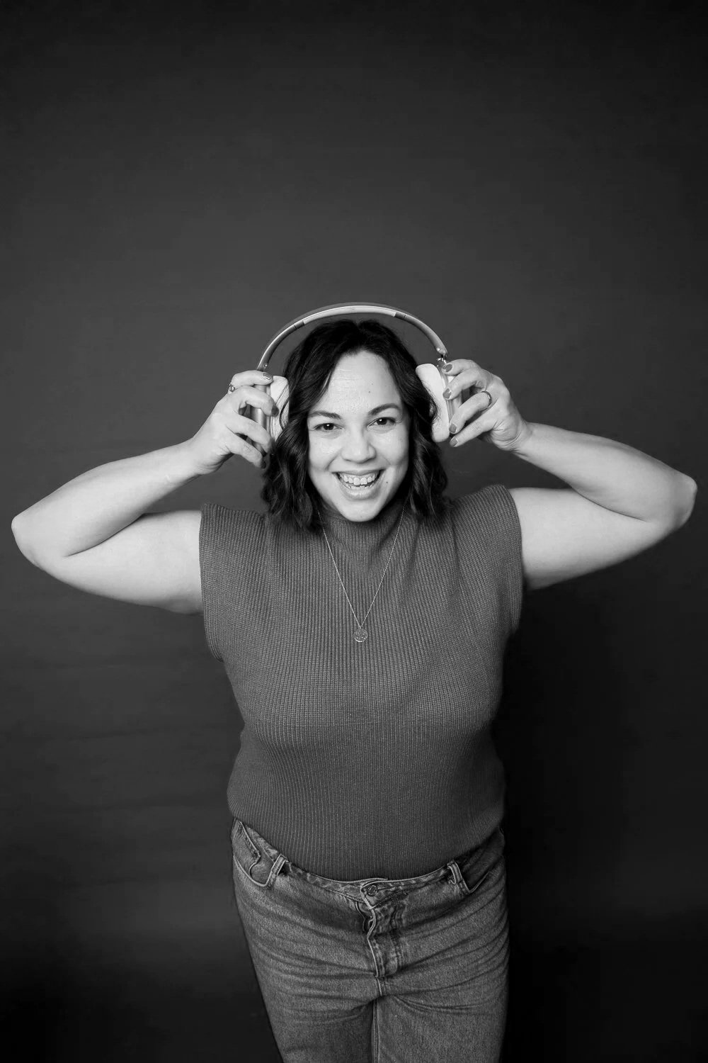 Joven mujer con gafas grandes y camiseta de punto, sonriendo y sosteniendo auriculares grandes. Fondo oscuro, en blanco y negro.