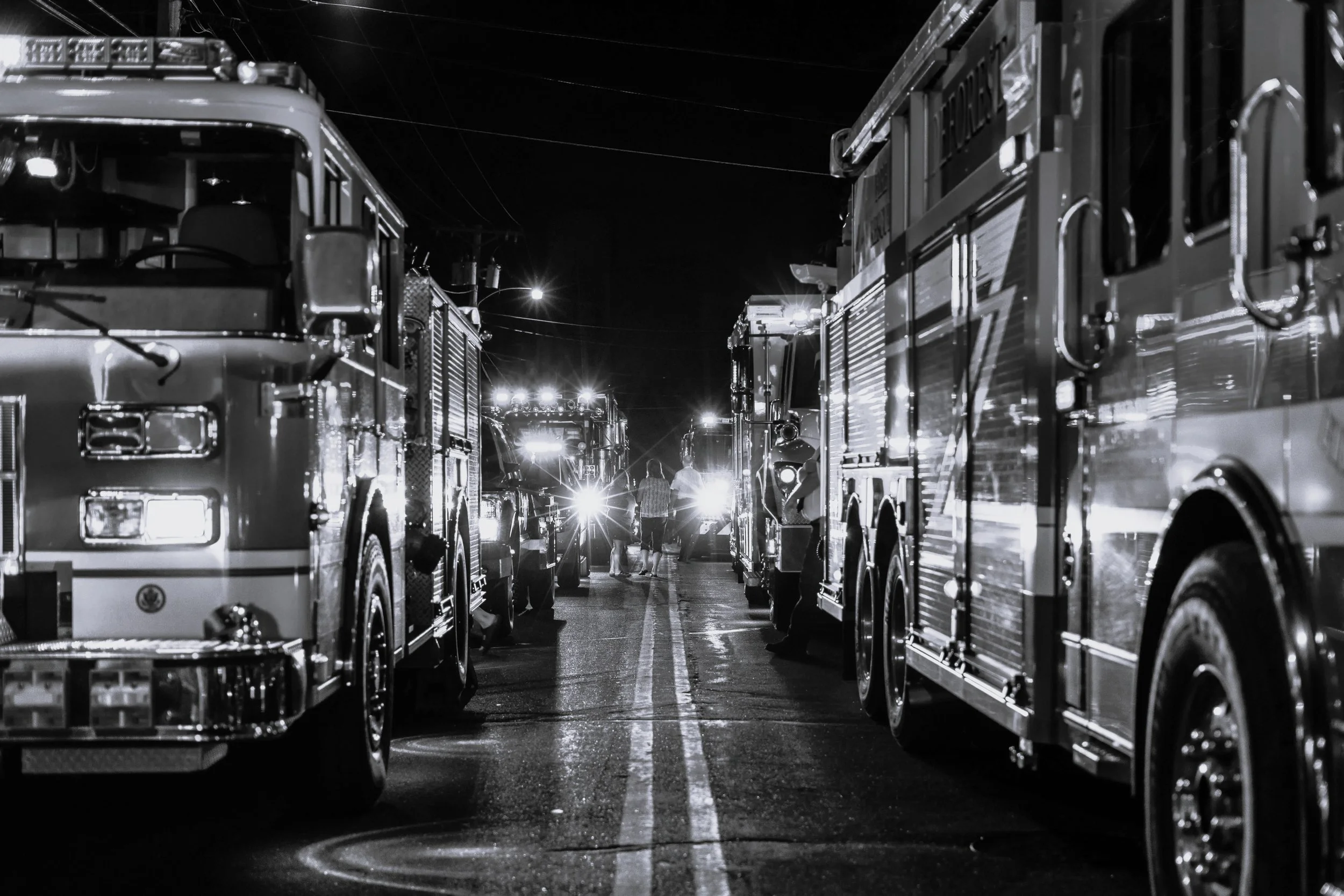 Nighttime scene of fire trucks and emergency vehicles parked closely together, with people walking between them under streetlights.