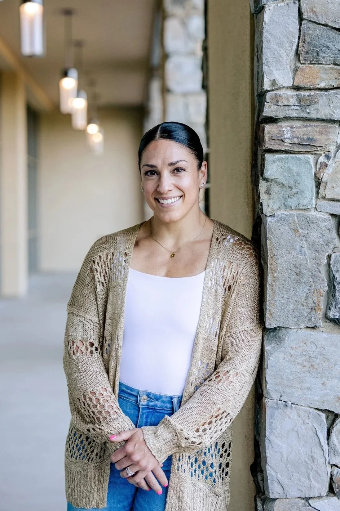 A woman with dark hair in a low bun, wearing a white top, a tan crocheted cardigan, and jeans, standing outdoors next to a stone wall and smiling.