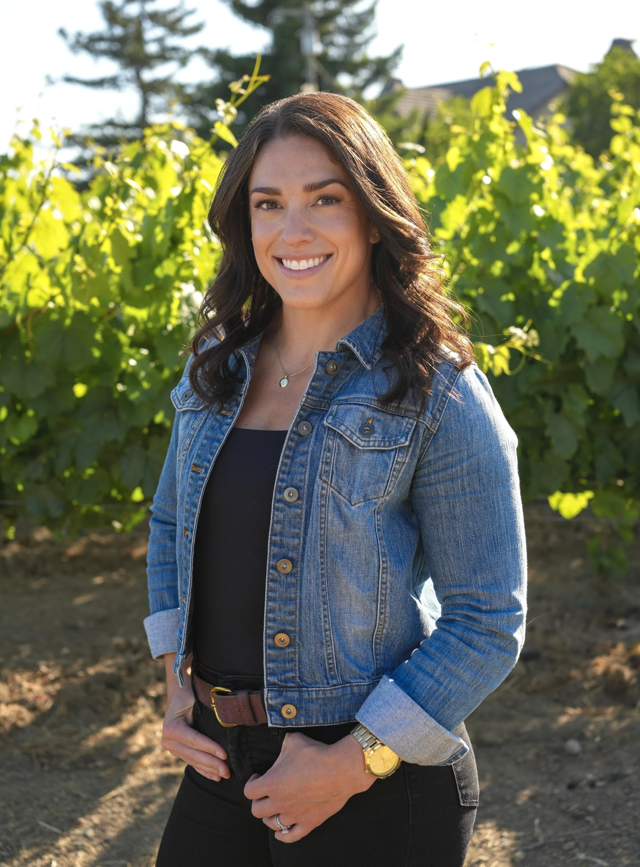 A woman with long dark hair smiling outdoors in a vineyard, wearing a denim jacket, black top, and a gold watch.