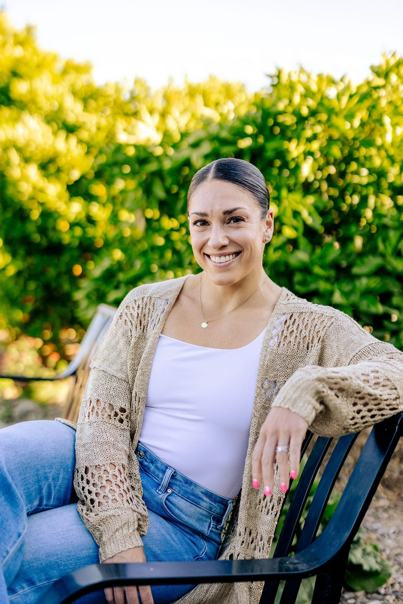 A woman smiling while sitting outdoors on a black metal chair. She is wearing a beige crocheted cardigan, white top, and blue jeans. There is green foliage in the background and sunlight.