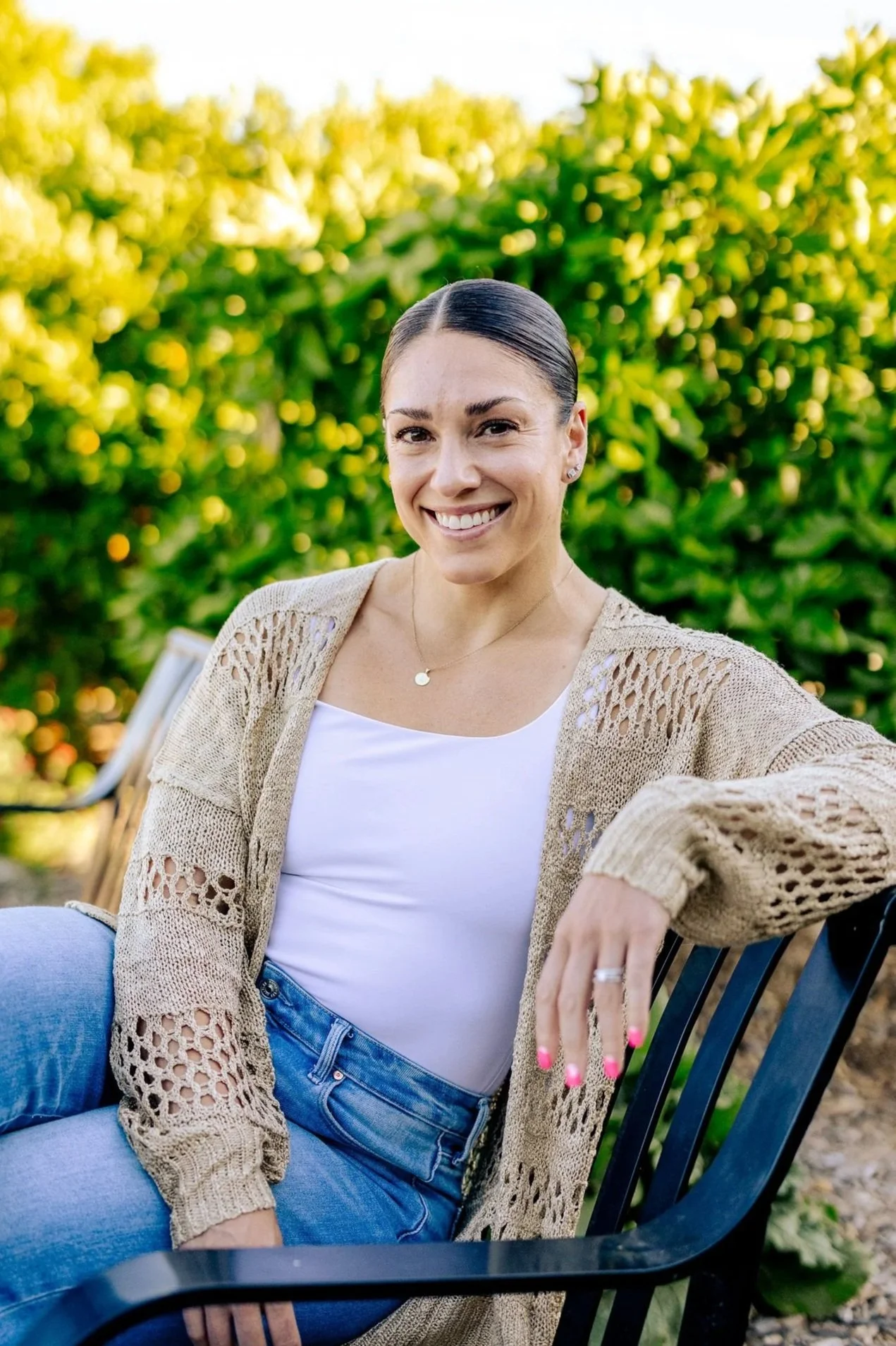 A woman with dark hair tied back, smiling, seated outdoors on a black metal chair with a leafy green background, wearing a beige knitted cardigan, white top, and blue jeans.
