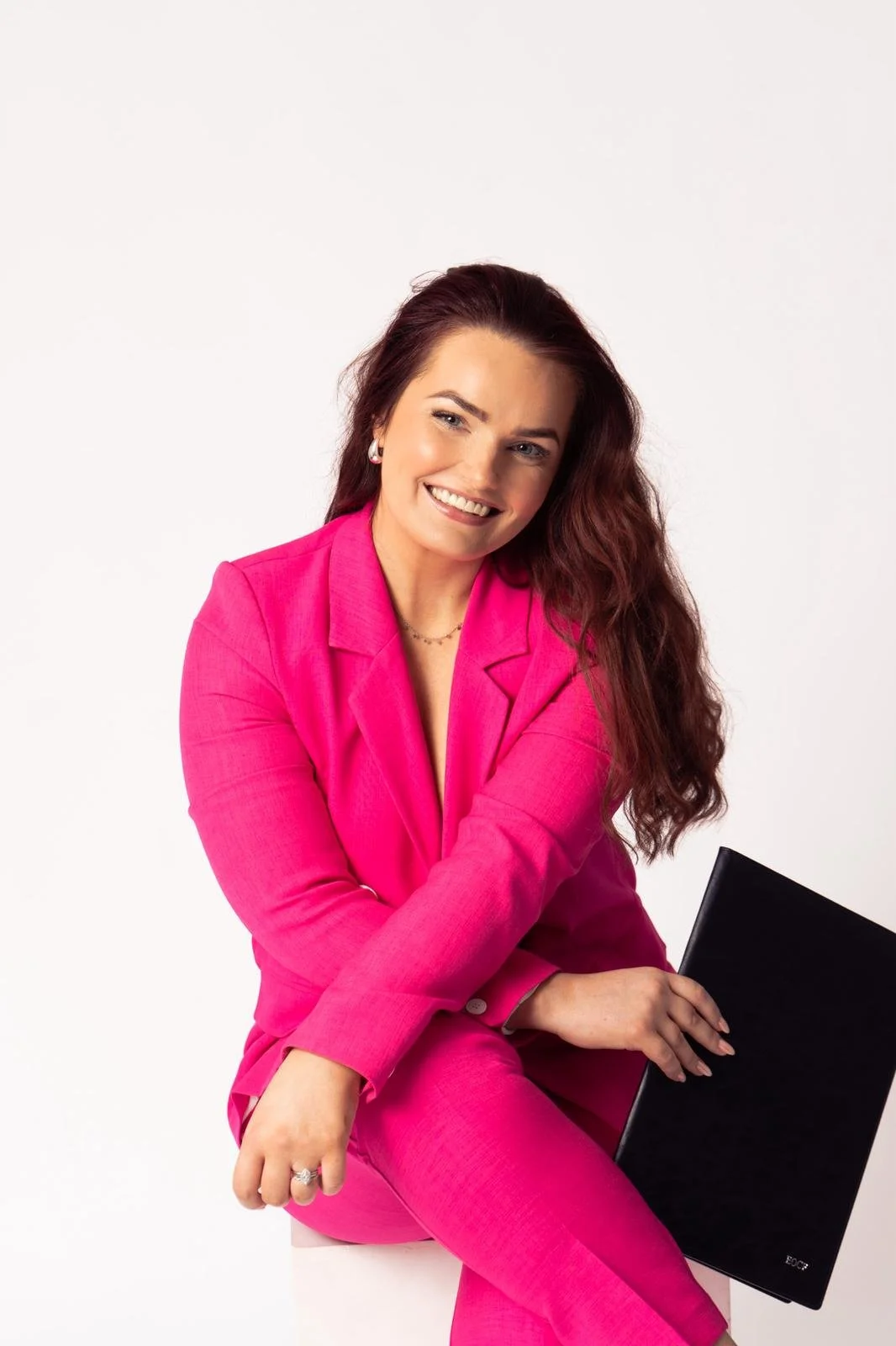 A woman with long, wavy auburn hair wearing a bright pink suit, sitting holding a black wedding celebrant folder on her lap, smiling against a white background.