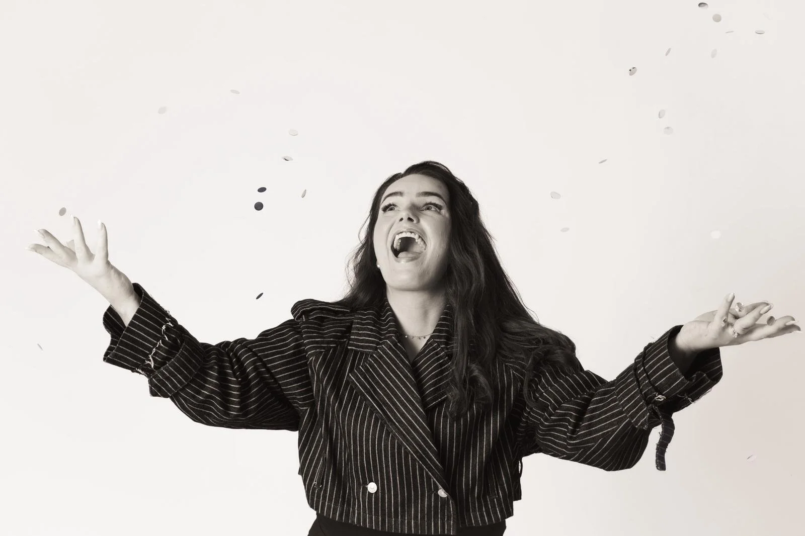 A woman with dark hair, wearing a pinstripe blazer, is celebrating with arms outstretched and confetti falling around her.