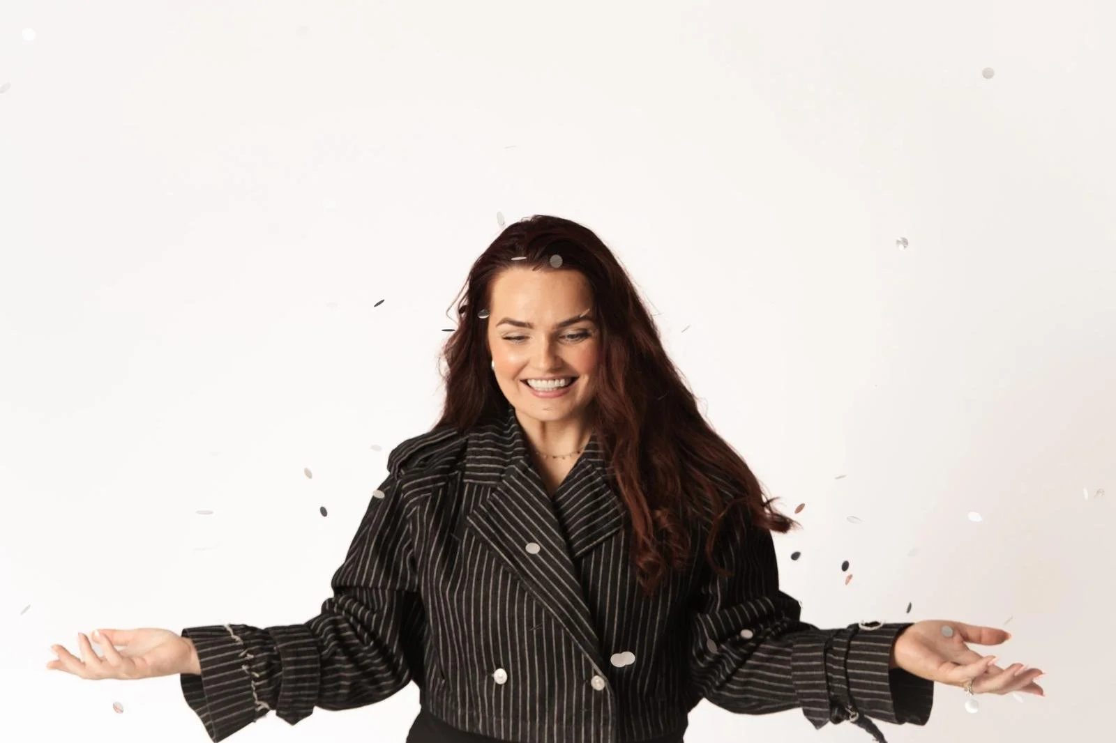 Smiling woman in a black pinstripe blazer with confetti falling around her against a white background.