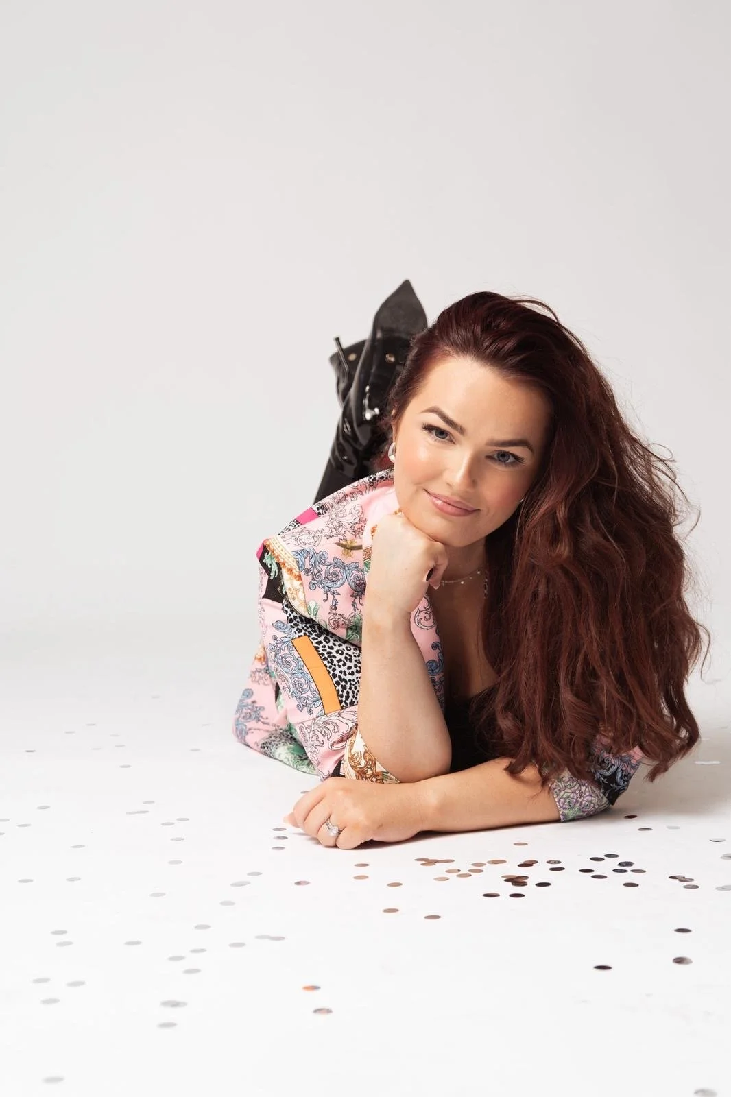 Woman with long, wavy red hair lying on a white surface surrounded by scattered gold and silver confetti, wearing a colorful patterned jacket and black boots, resting her chin on her hand and smiling.
