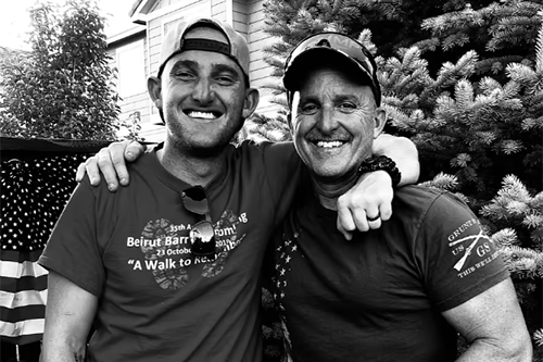 Two men smiling outdoors, with one man having his arm around the other's shoulder, in front of a Christmas tree and house, in black and white.