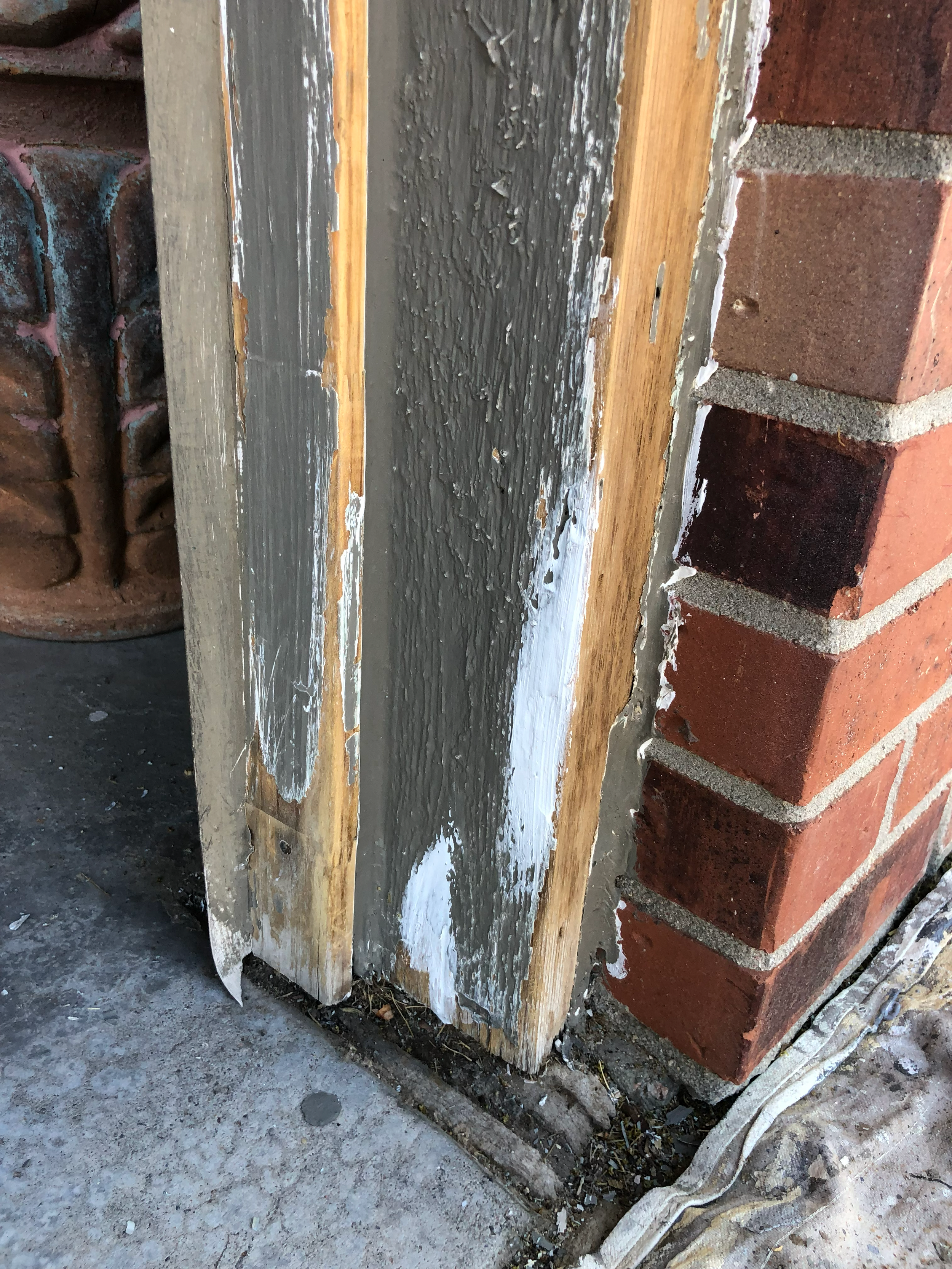 Close-up view of a wooden beam with partially painted or coated gray and white surface, adjacent to a brick wall and a concrete sidewalk.