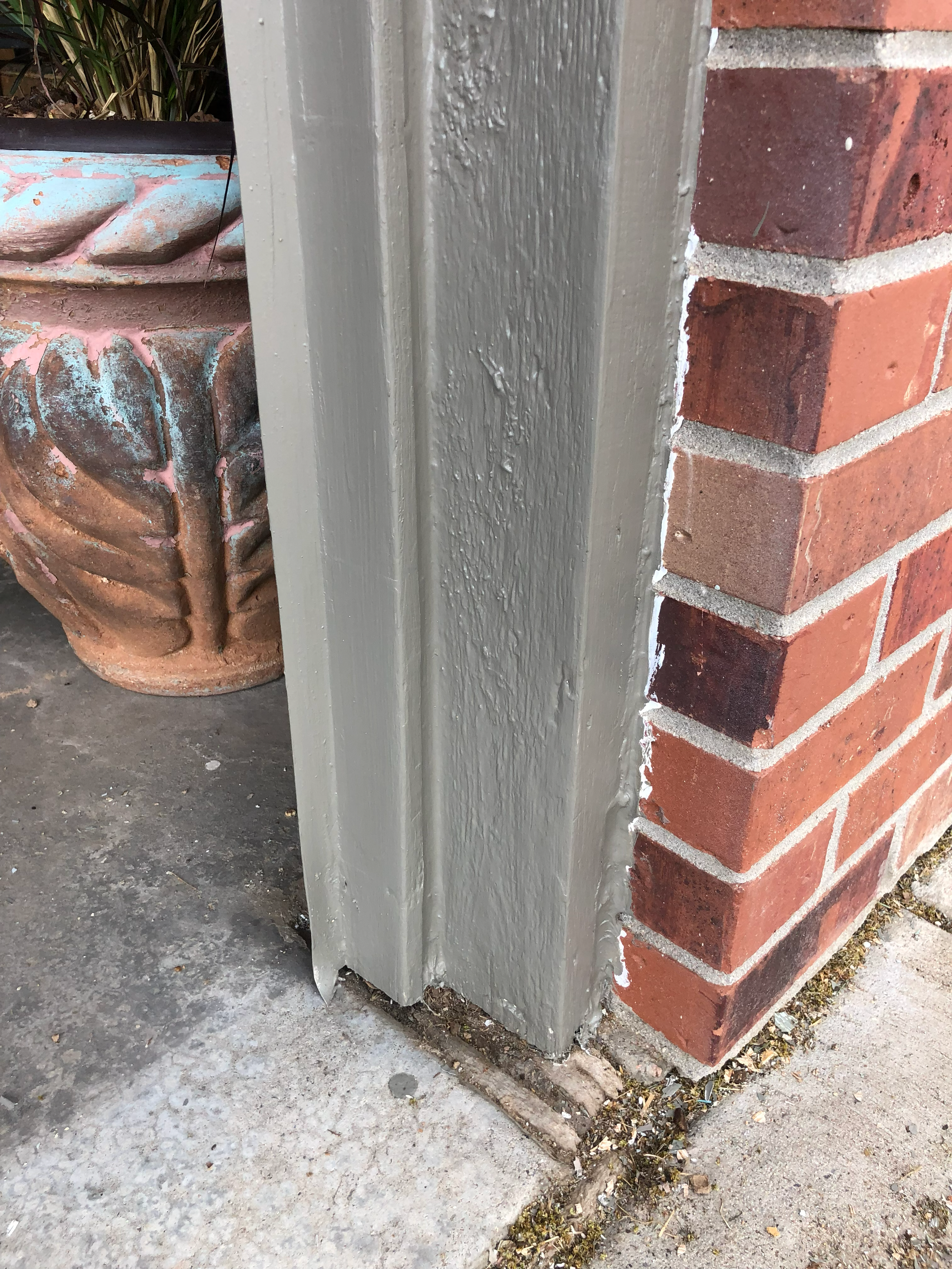 Close-up of a corner of a brick building with a wooden post painted gray and a decorative clay pot nearby, on a concrete surface with some leaves and debris.