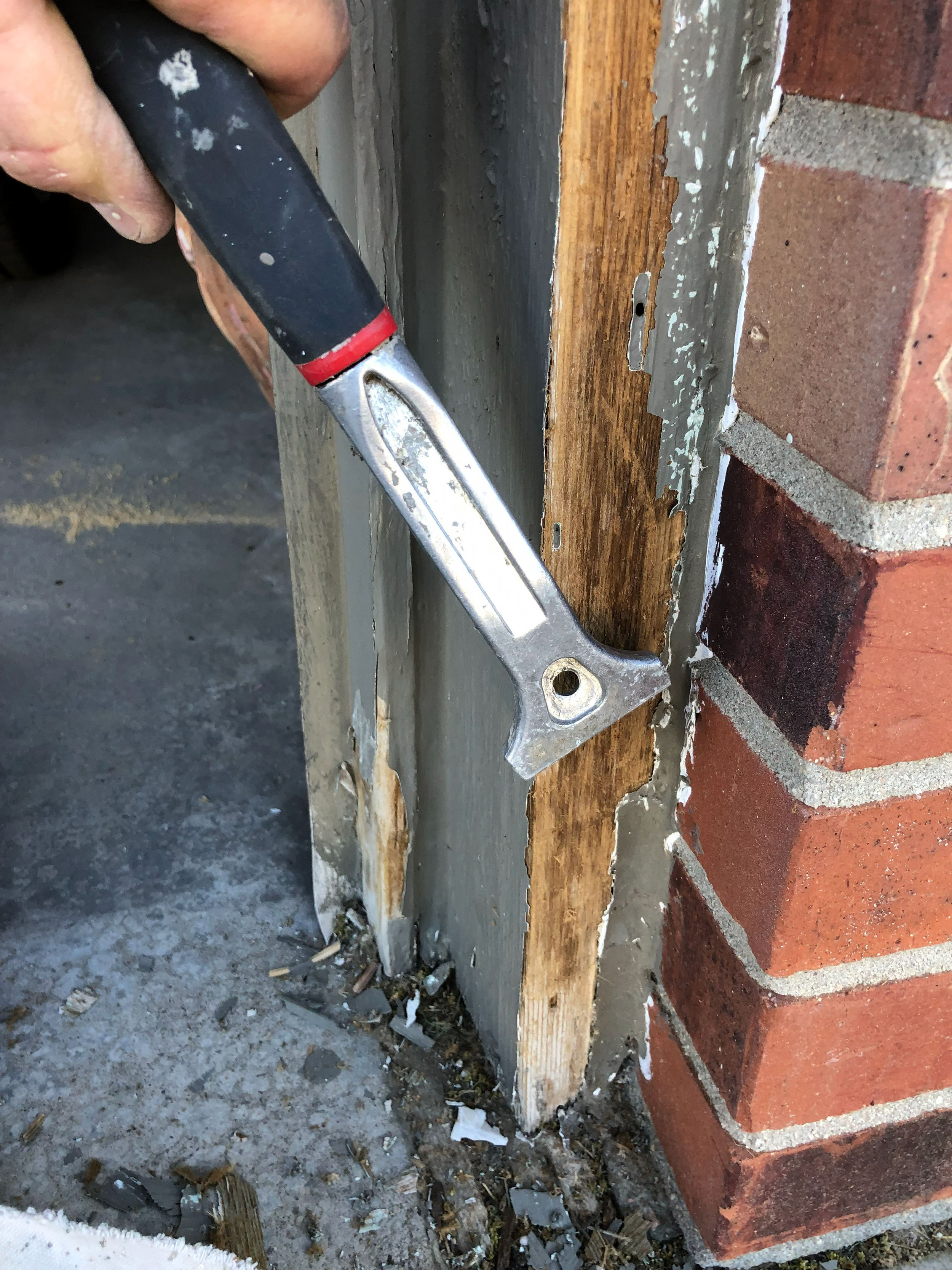 A hand holding a crowbar is prying off old wooden trim from a brick wall. The wall features a combination of brick and concrete, and the wooden trim shows signs of wear. Debris is scattered on the ground.