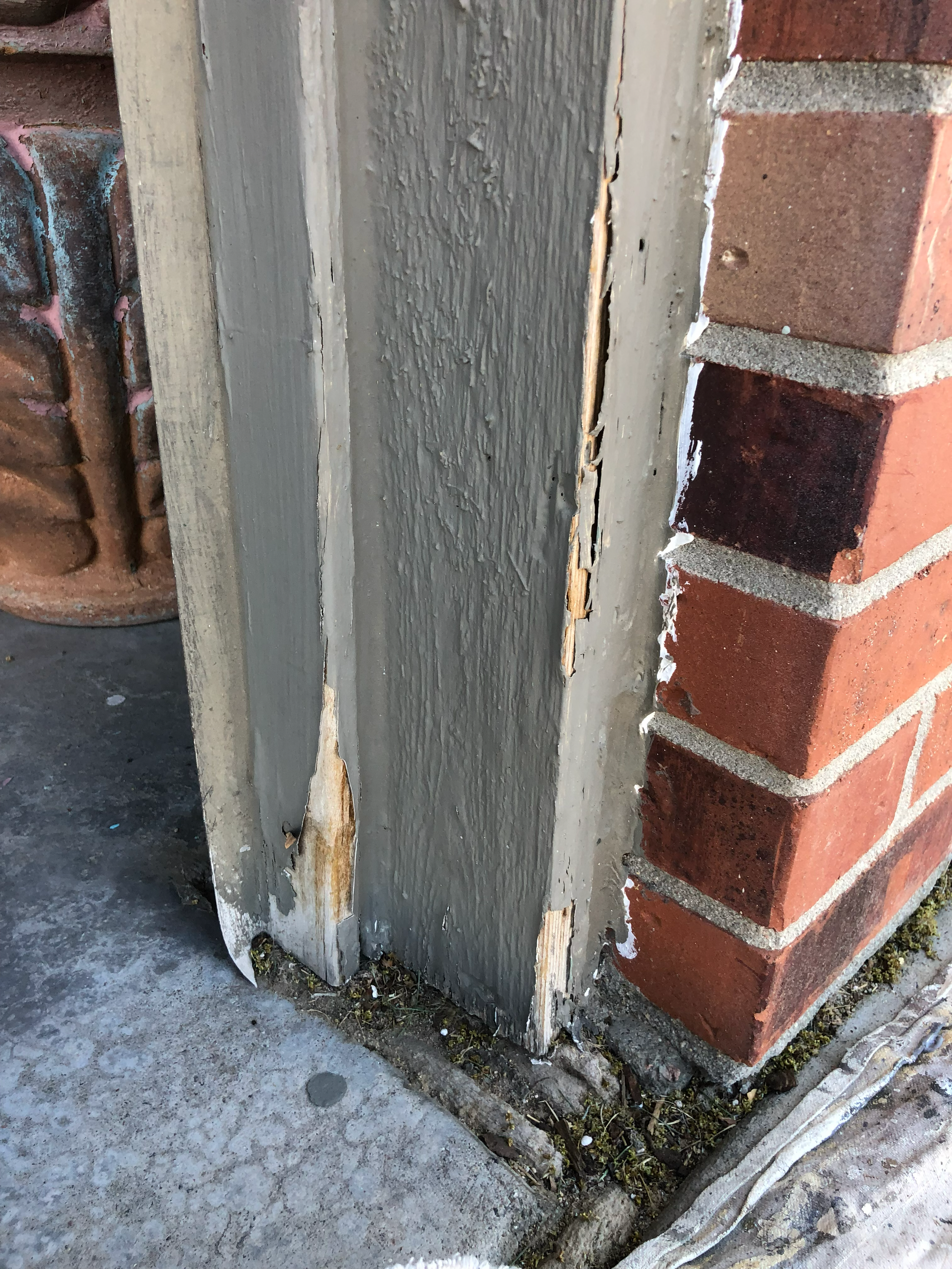 Close-up of a brick wall corner with a wooden board showing peeling paint and damage, on a cracked concrete surface with weeds growing at the base.