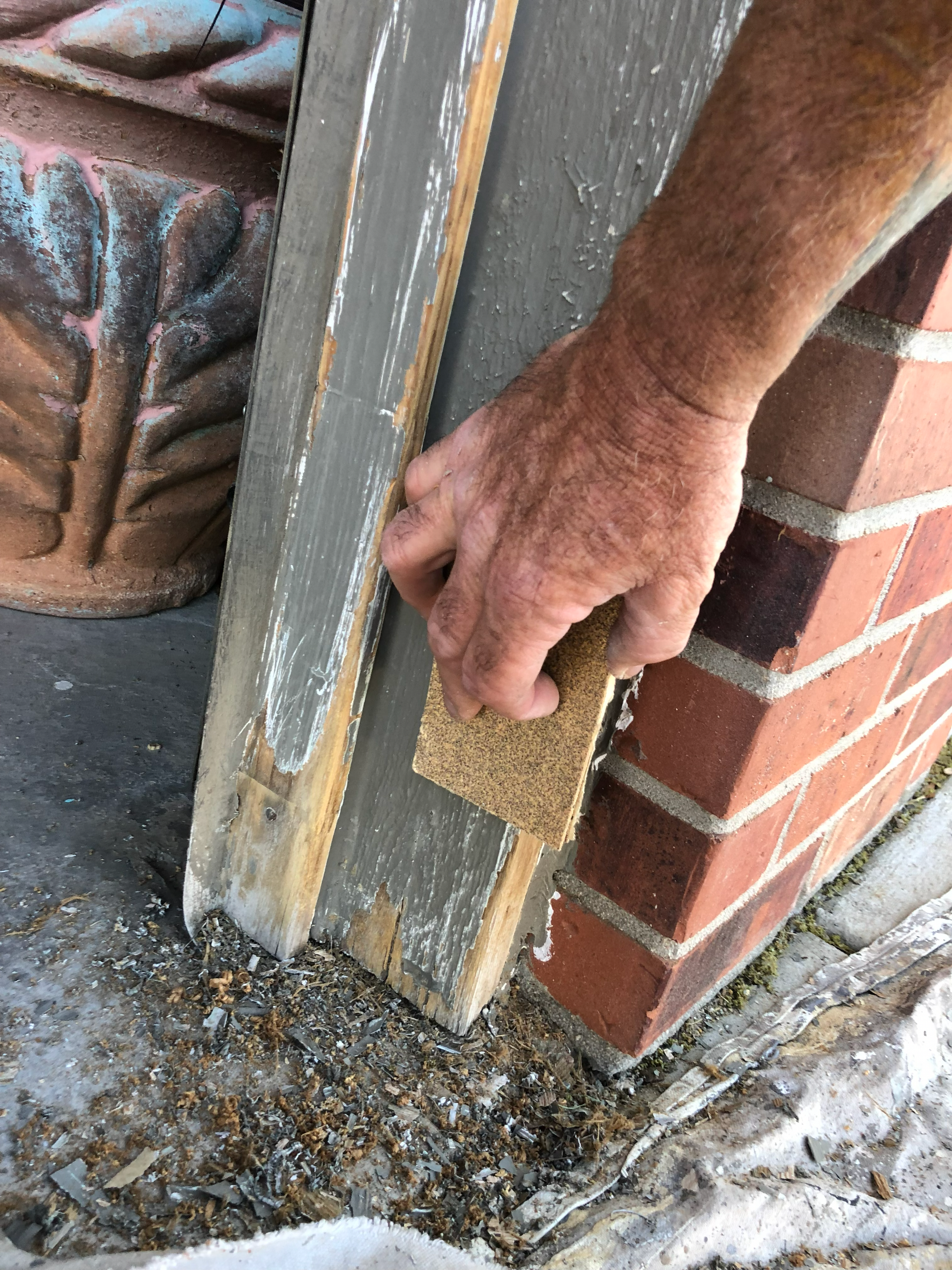 A person with a weathered hand is working with a paintbrush on a rough wooden surface near a brick wall, with debris and dirt on the ground.