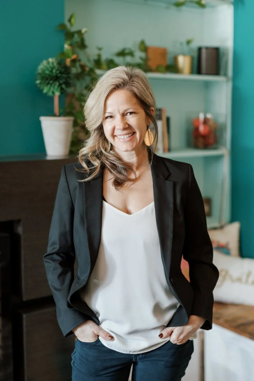 Woman smiling, standing in a cozy room with teal walls, a bookshelf, books, plants, and decor in the background.