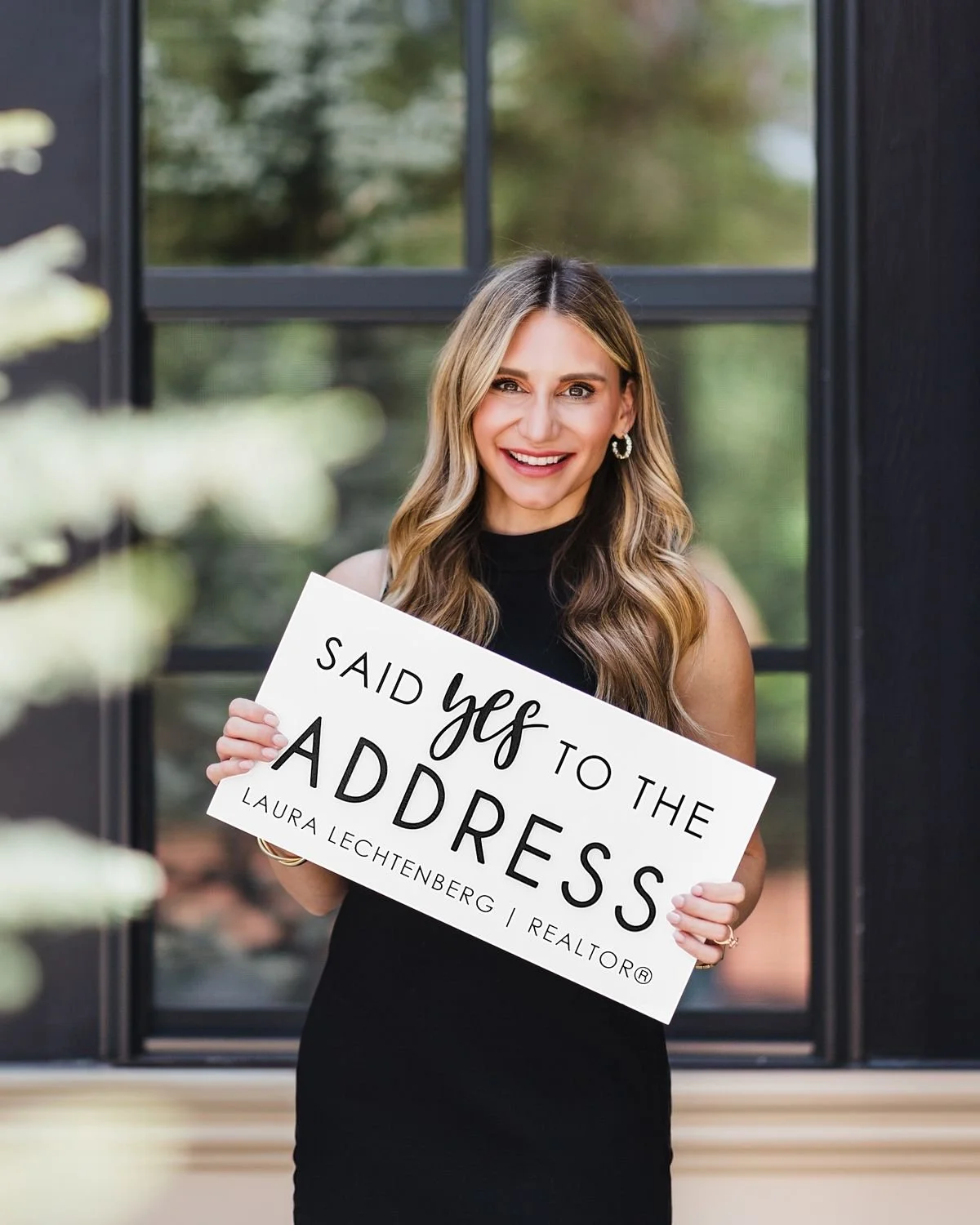 A woman with long, wavy hair smiling and holding a sign that reads 'Said yes to the address' in front of a large window with greenery outside.