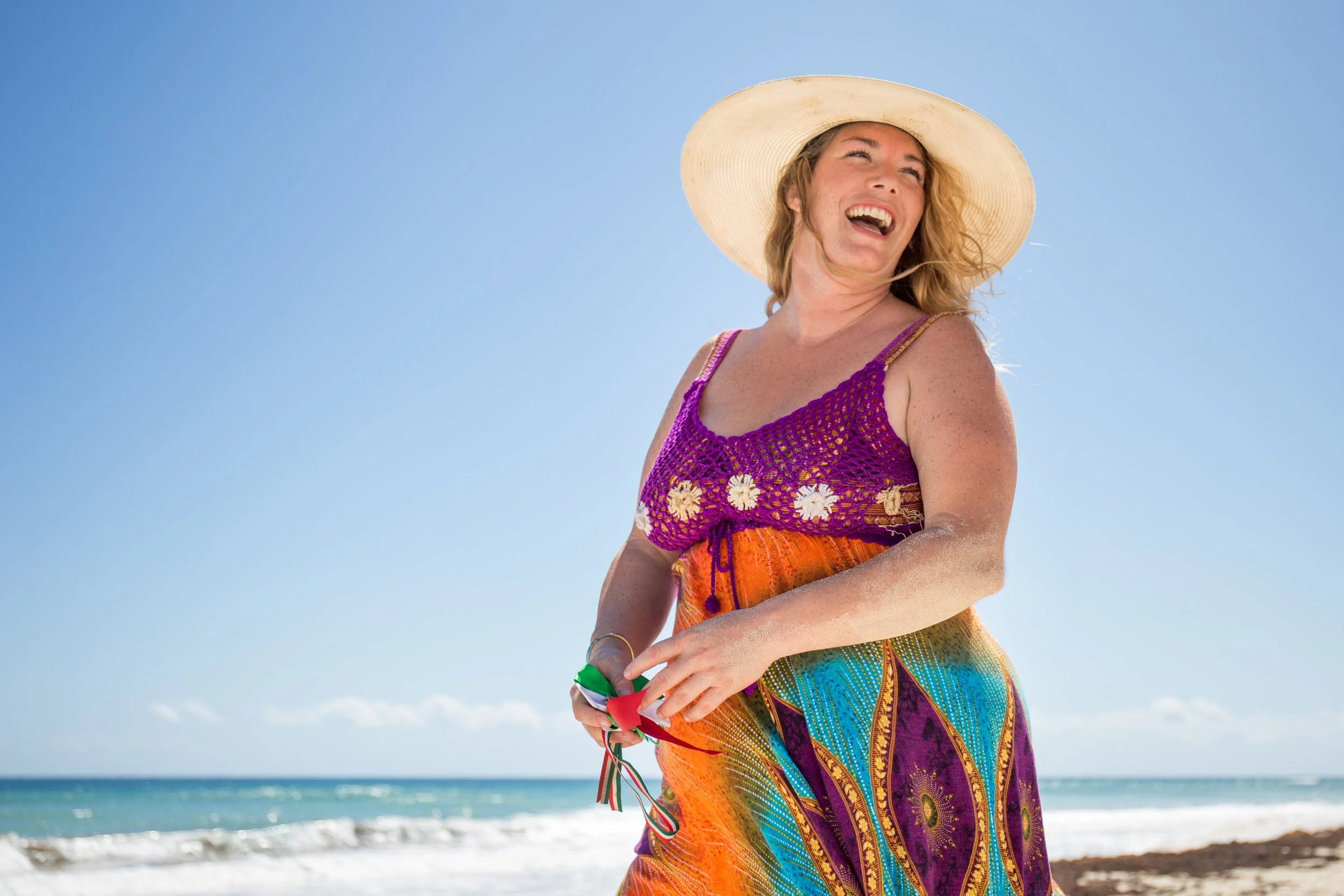 Woman smiling and enjoying the beach, wearing a wide-brimmed hat and a colorful, patterned dress, with the ocean and clear blue sky in the background.