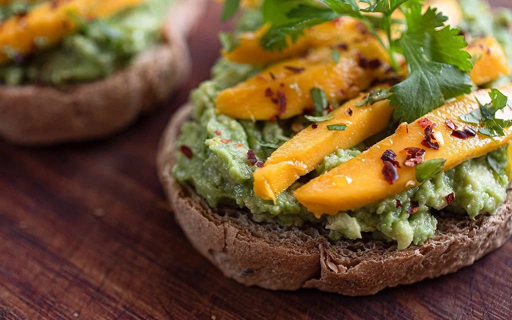 Close-up of an avocado toast topped with grilled mango slices, chili flakes, and cilantro on whole grain bread.