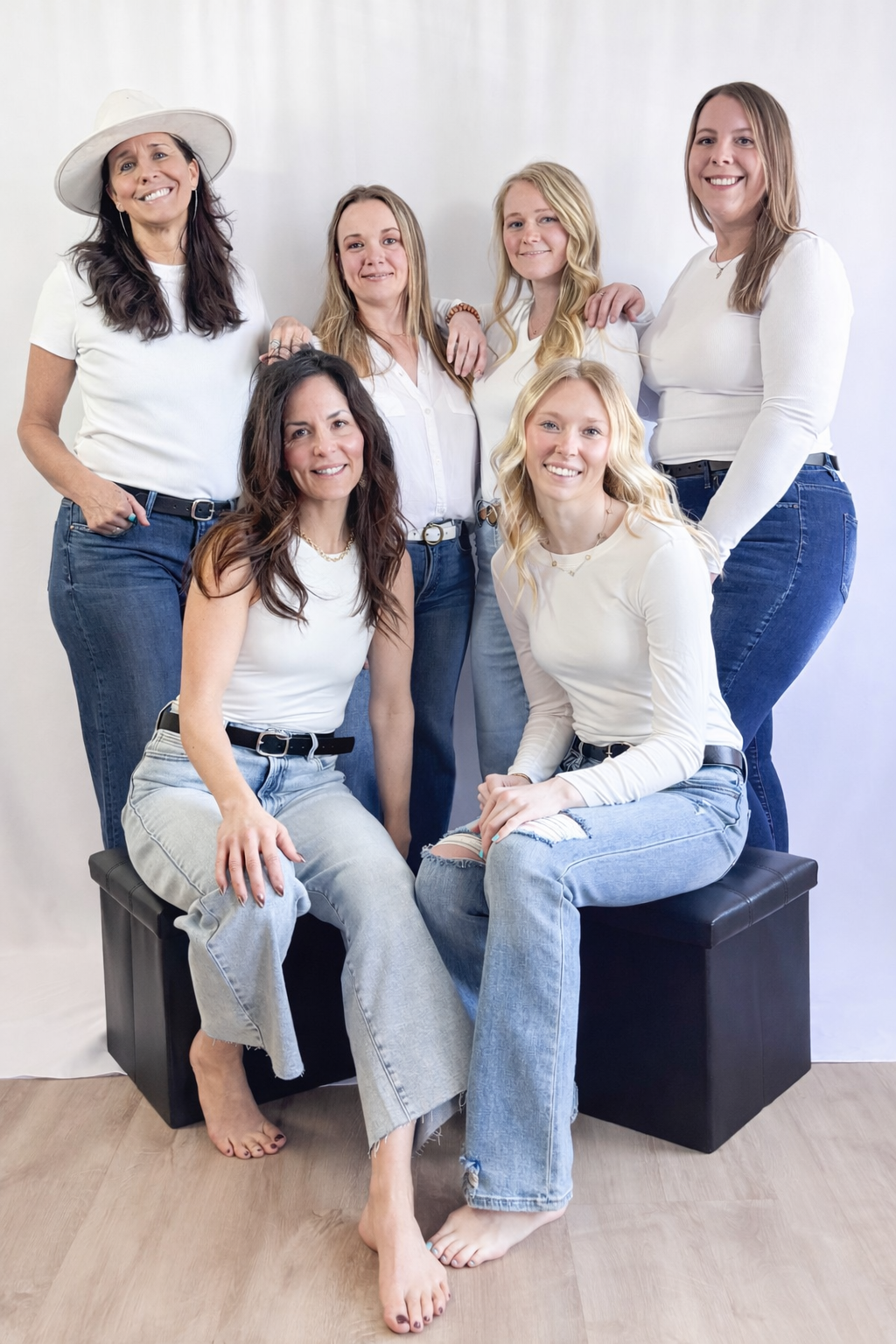 Group of seven women posing together, some sitting on black cubes and others standing against a white background, all smiling and dressed in casual white tops and jeans.