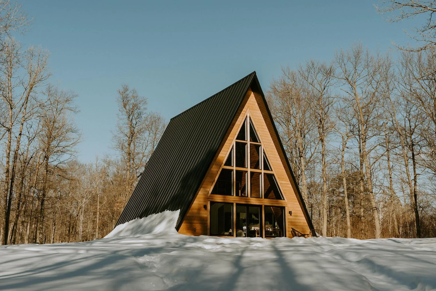 A modern A-frame cabin with large windows, surrounded by snow and a forest of leafless trees under a clear blue sky.