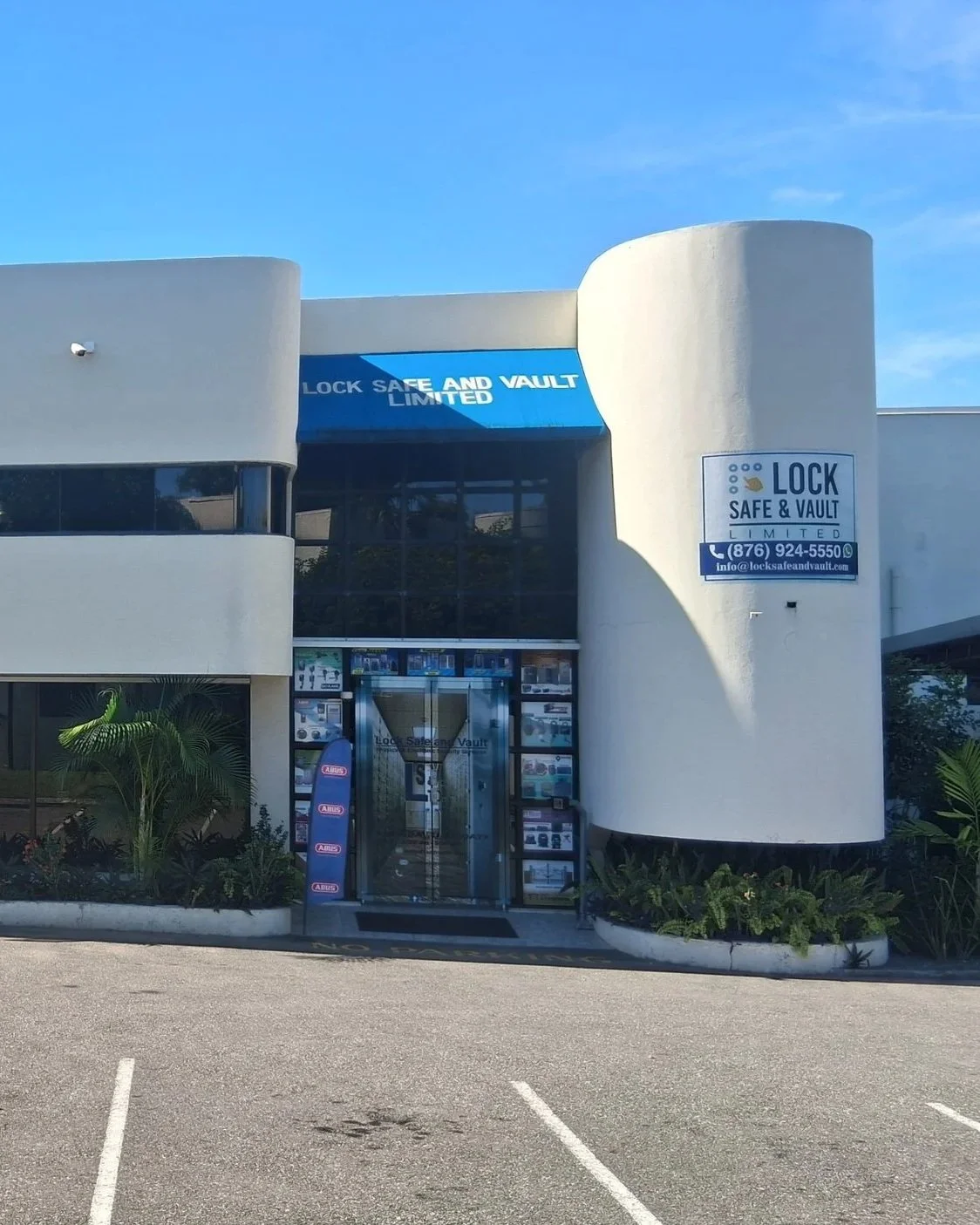 Front view of a white building with a blue awning that says "LOCK SAFE AND VAULT LIMITED" and a sign on the right side with the same name, phone number, and email. There are plants near the entrance and parking spaces in front.